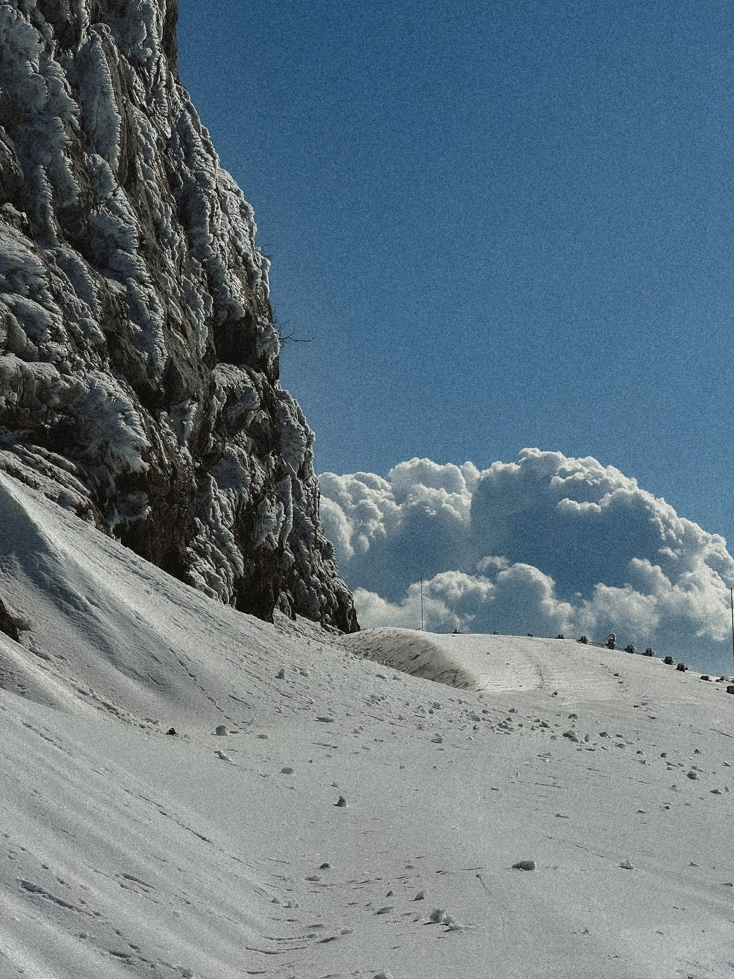 Snow-covered mountain slope with a rocky cliff on the left and a clear blue sky with large white clouds in the background.