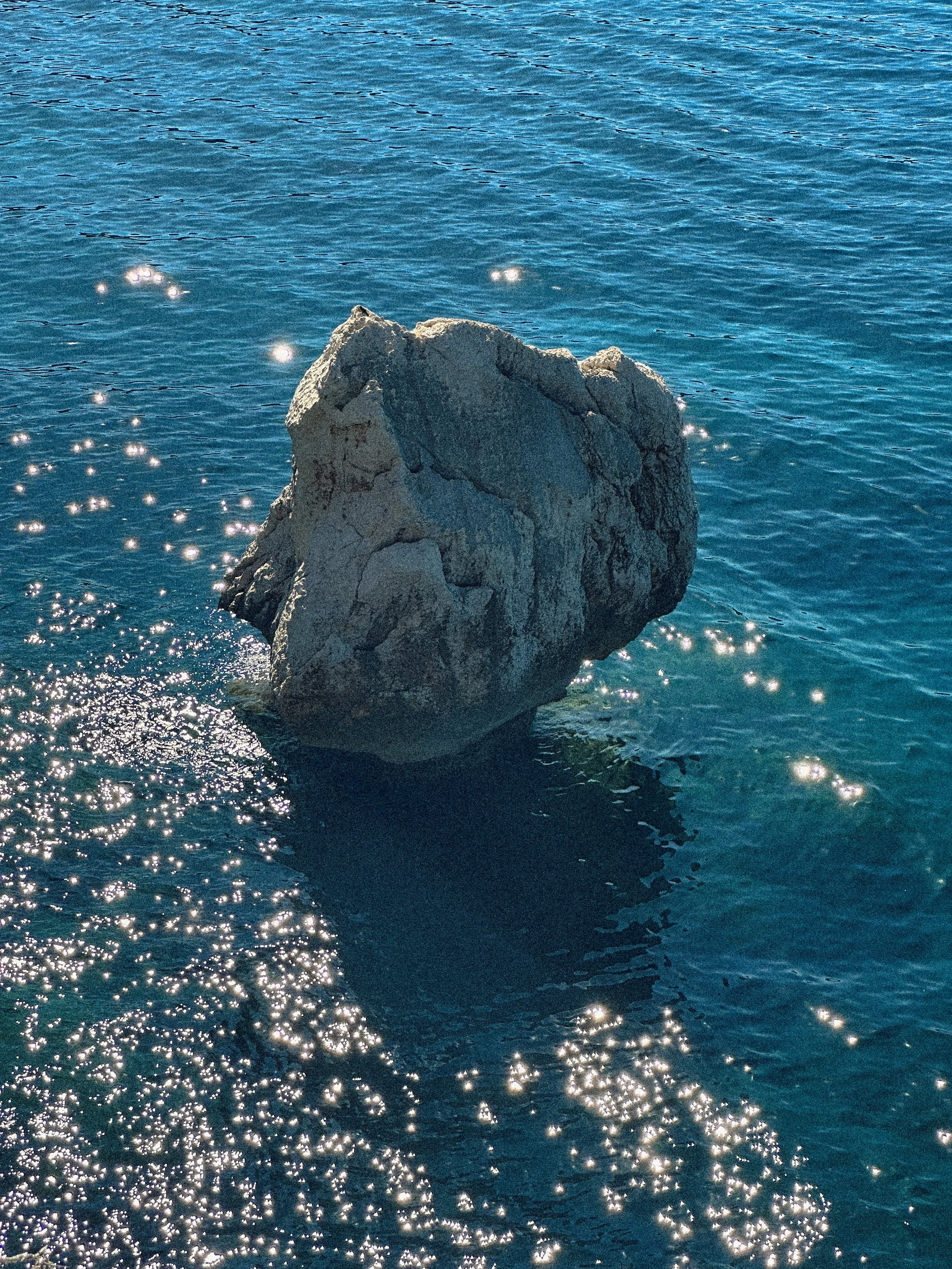 A large rock in the water with sunlight creating sparkling reflections on the surface.