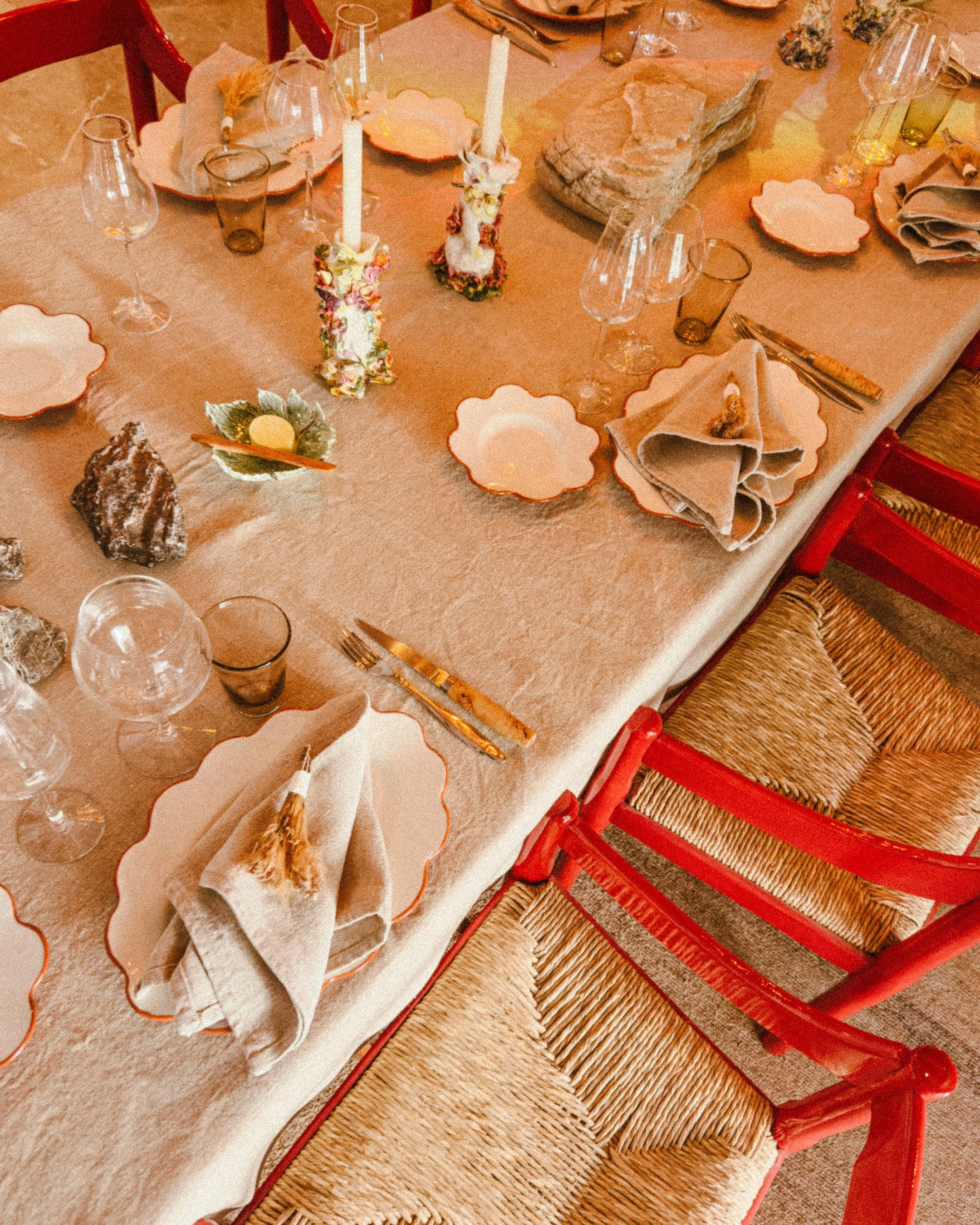 A decorated dinner table with beige linens, orange-trimmed plates, beige napkins with a tassel, silverware, wine glasses, candle holders with candles, and various rocks and decorative items.