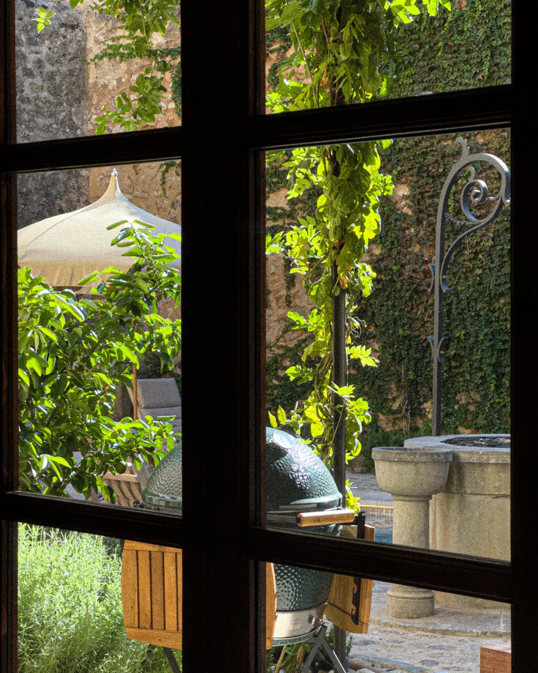 A view through a window shows a backyard with greenery, a large umbrella, a green barbecue grill, and a stone water fountain.