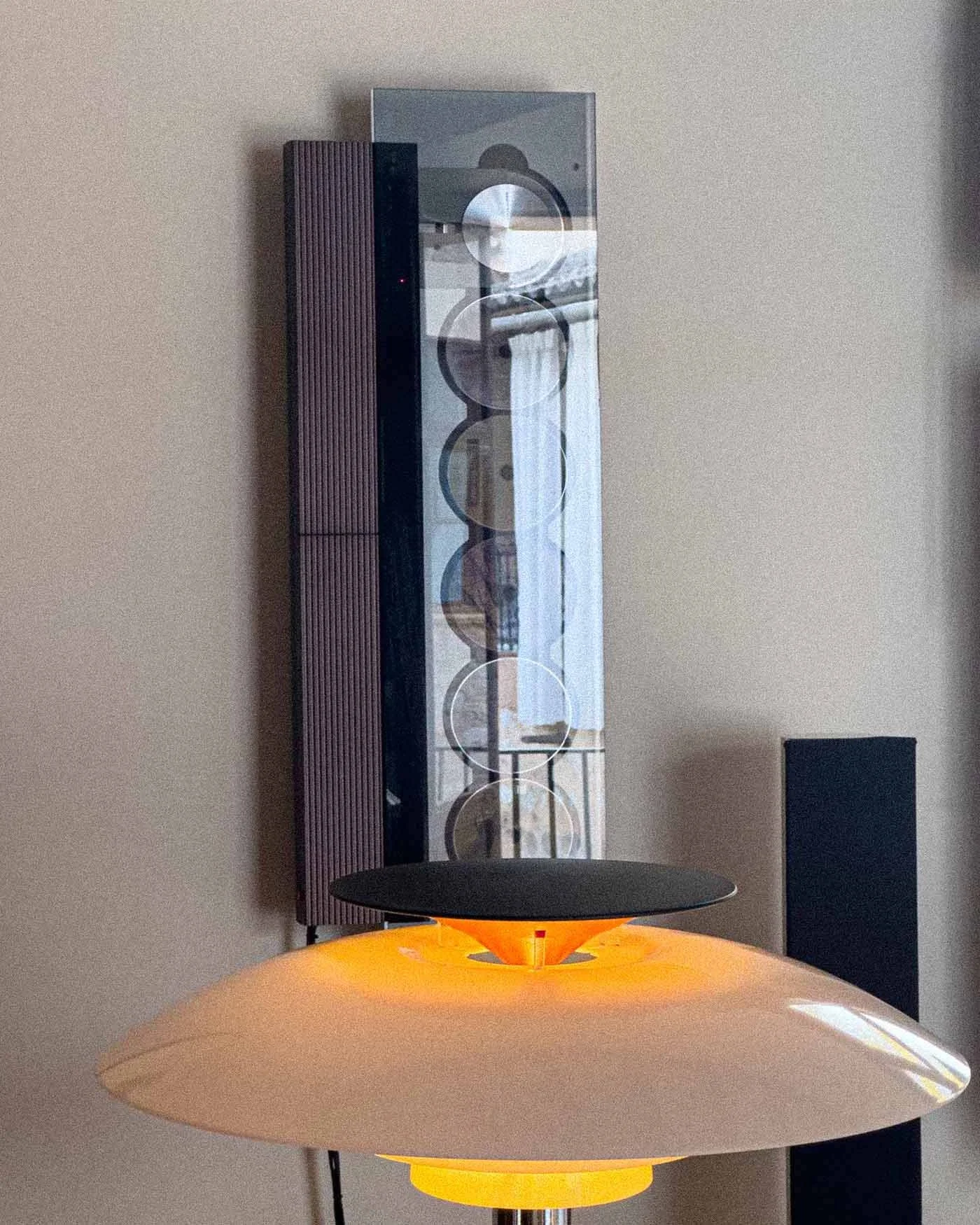 A modern ceiling lamp with a black top and a white, rounded, disc-shaped bottom, illuminated with warm light, with a wall-mounted speaker and a glass display case with multiple round objects behind it.