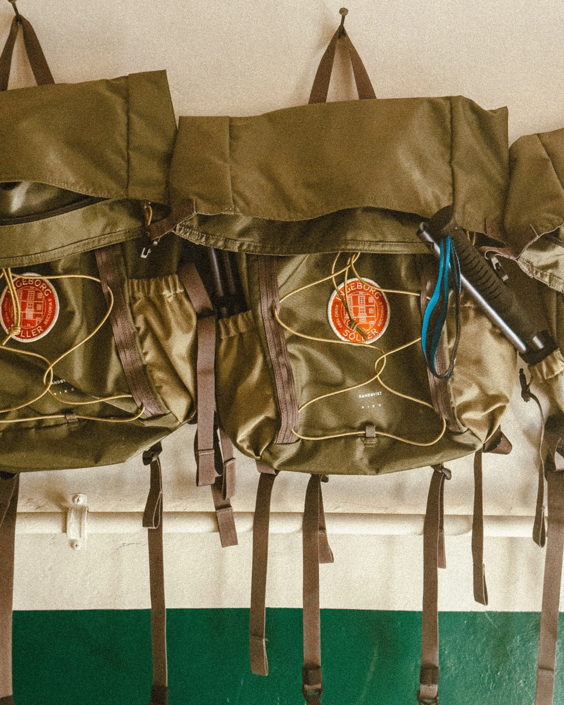 Three olive-green hiking backpacks with brown straps hanging on a wall, with a blue and black pen attached to one of the backpacks.