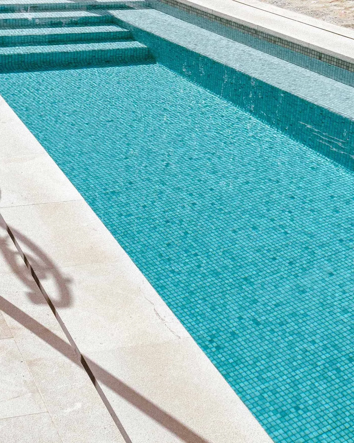 Close-up of a swimming pool with small blue mosaic tiles and steps leading into the water, surrounded by a white concrete deck.