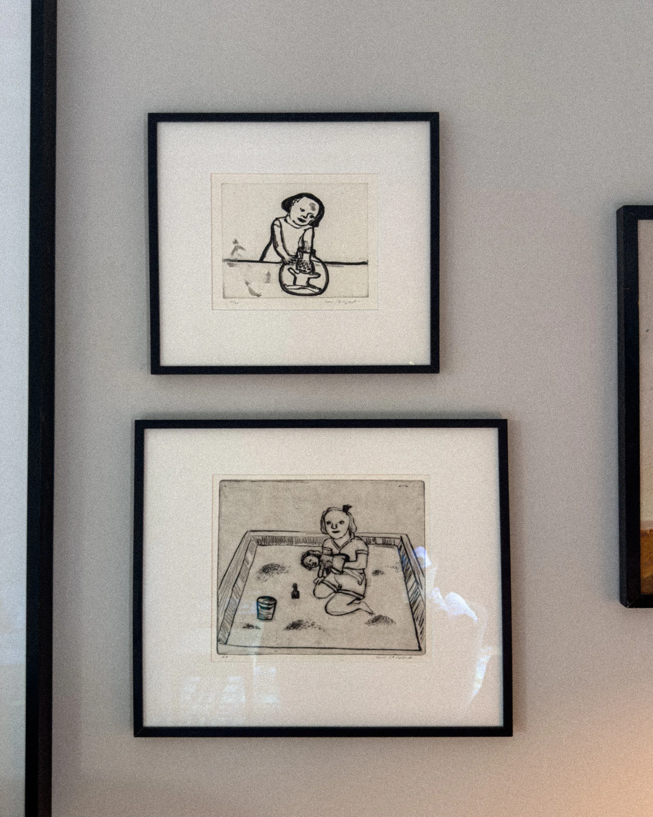 Two framed black and white drawings of children, one above the other, hanging on a wall. The top drawing depicts a girl with short hair holding an object in her hand, and the bottom shows a girl inside a sandbox holding a toy, with sand and toys around her.