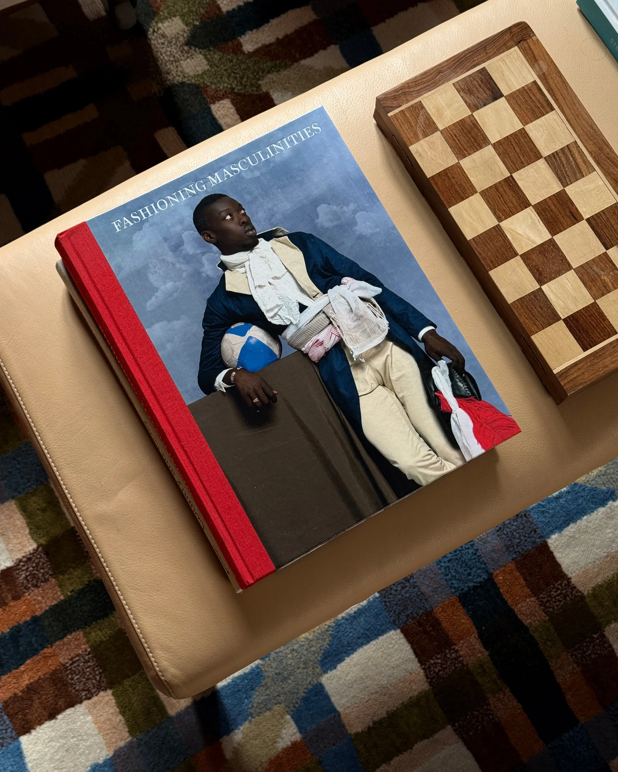 A book titled 'Fashioning Masculinities' lies on a beige leather surface, with a man in historical attire holding a ball and a bag on the cover. Next to it is a wooden chessboard.