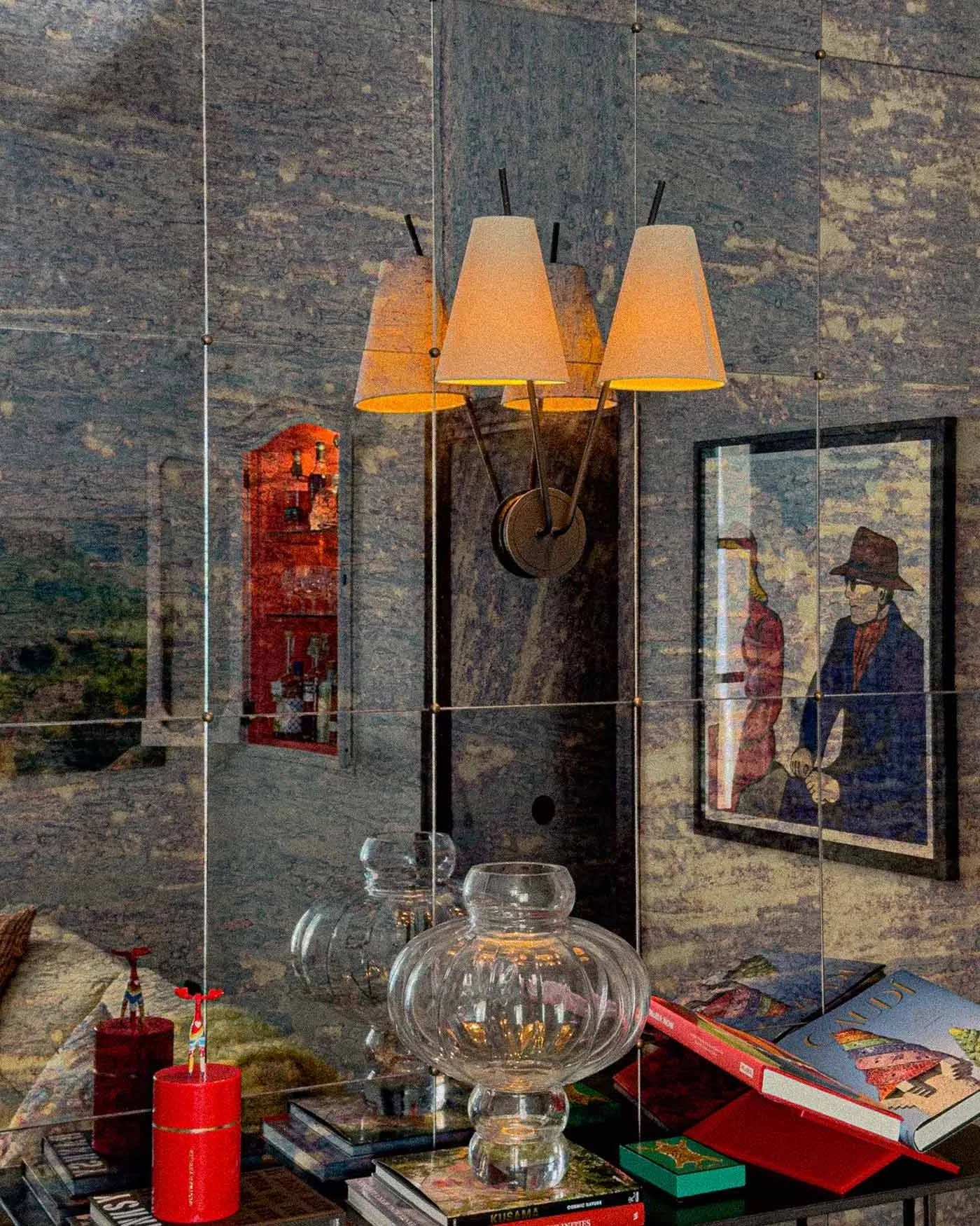 Interior view of a room with a stone wall, a modern wall lamp with three beige lampshades, framed artwork, glass vases, books, and a red candle on a table.