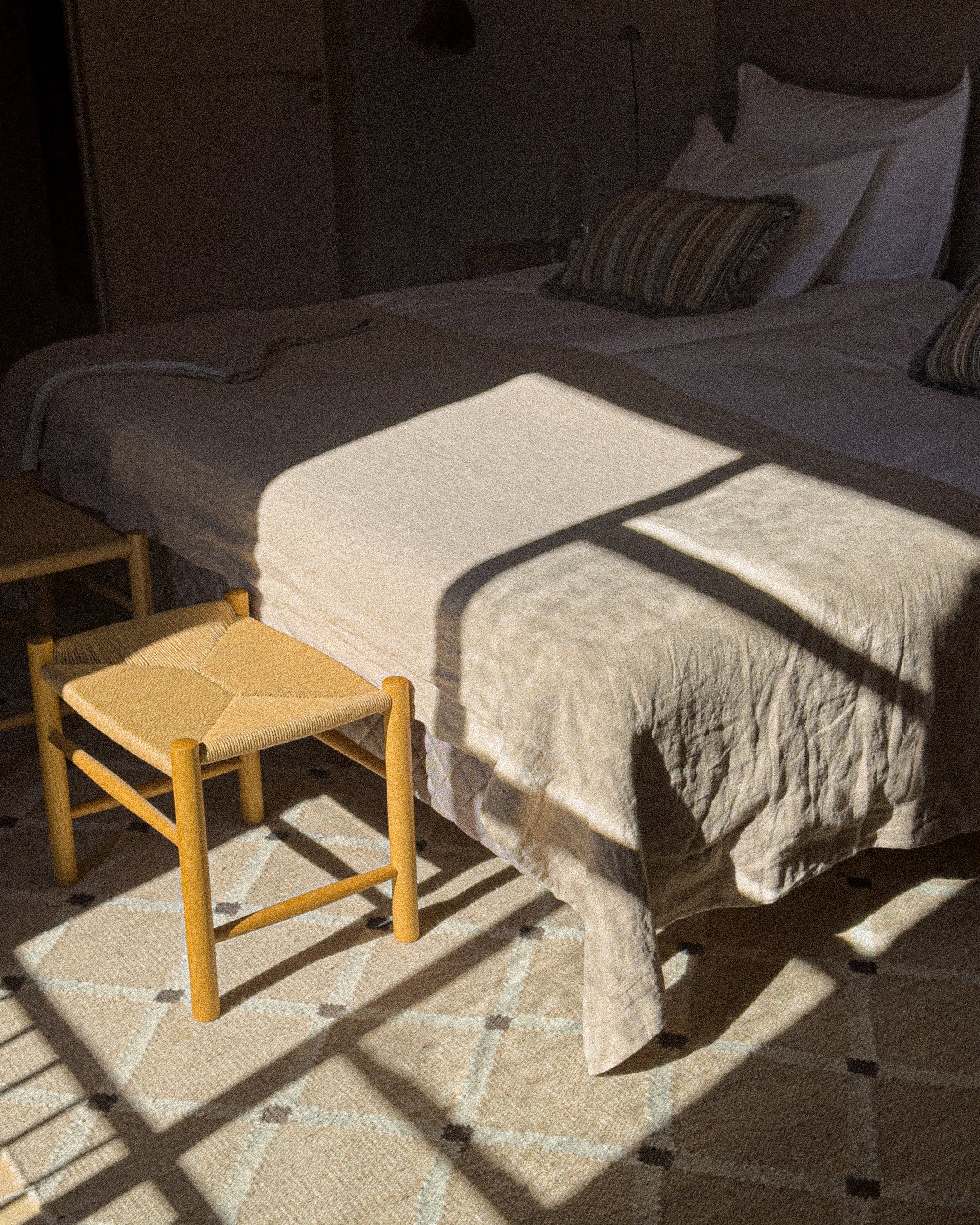 A bed with white bedding and striped pillows, sunlight casting shadows through a window onto the bed and beige patterned rug, a small wooden stool beside the bed, and a nightstand with a lamp in the background.