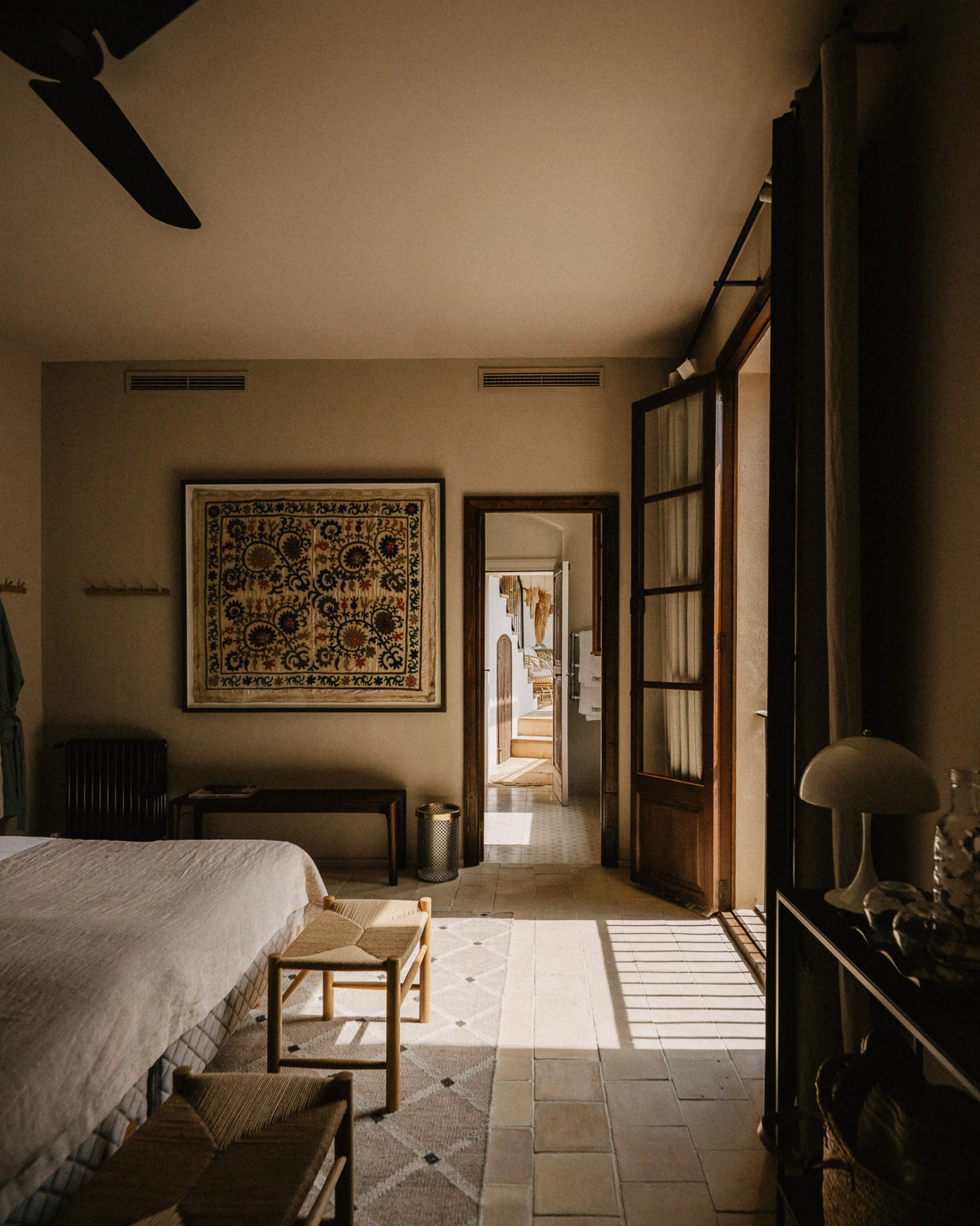 Hotel room with a bed, two small chairs, a rug, a large framed artwork, a mirror, and an open door leading to a hallway with stairs.