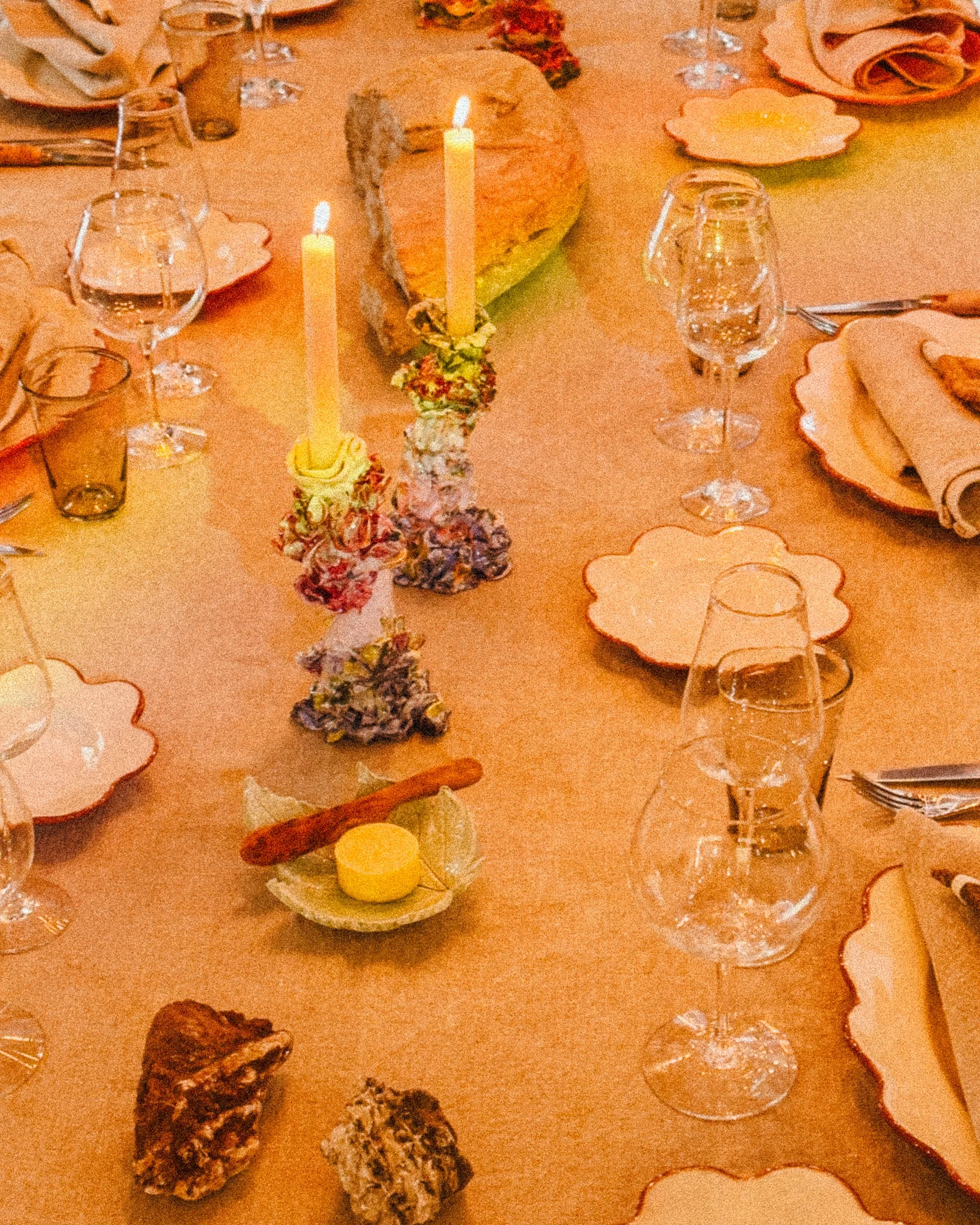 A dinner table decorated with two lit candles, a large bread loaf, assorted nuts, and various dishware, including wine glasses, plates, and cutlery, set for a formal meal.