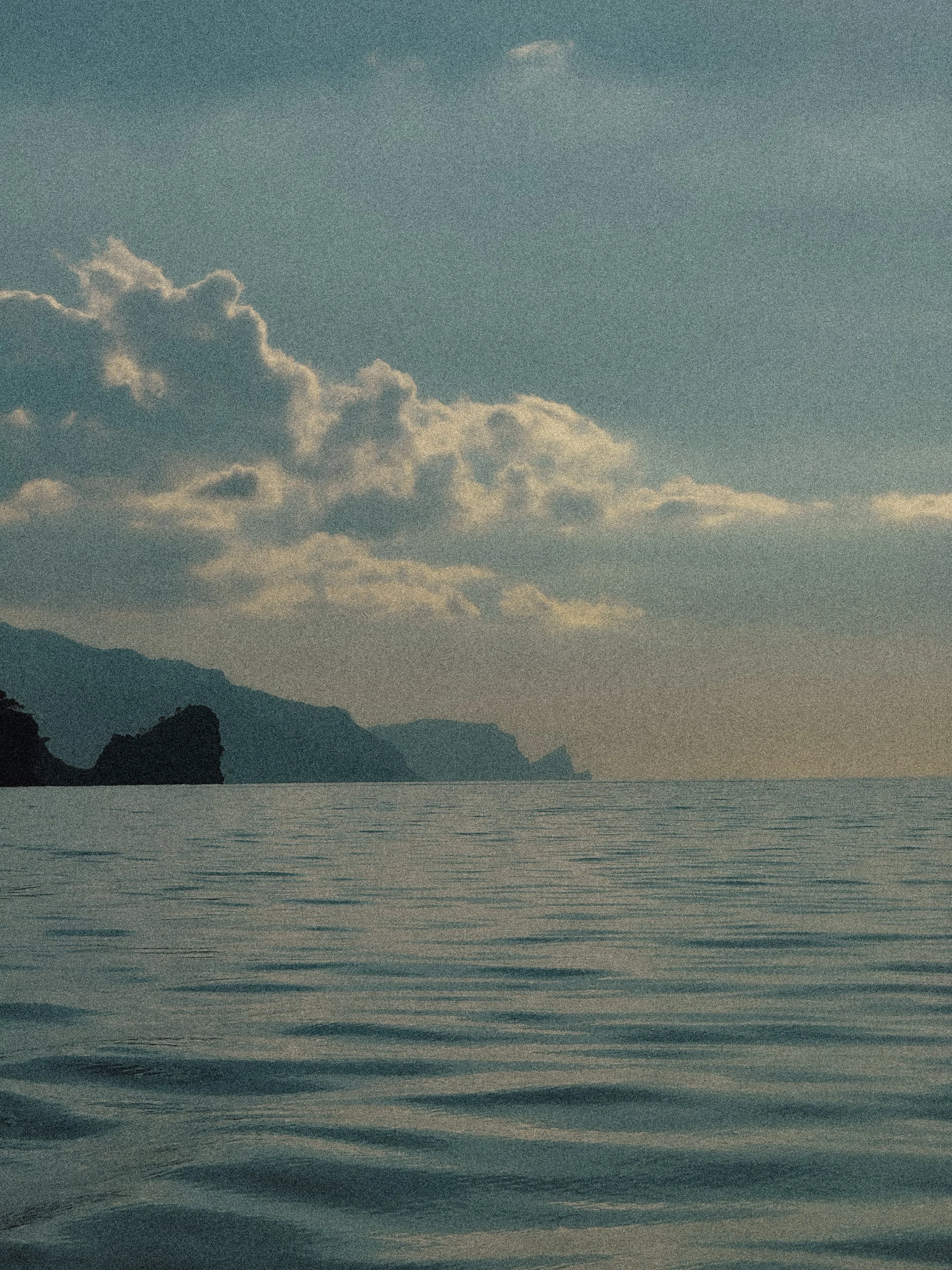 Calm sea with gentle waves, distant coastline with cliffs and mountains, partly cloudy sky with some sunlight.