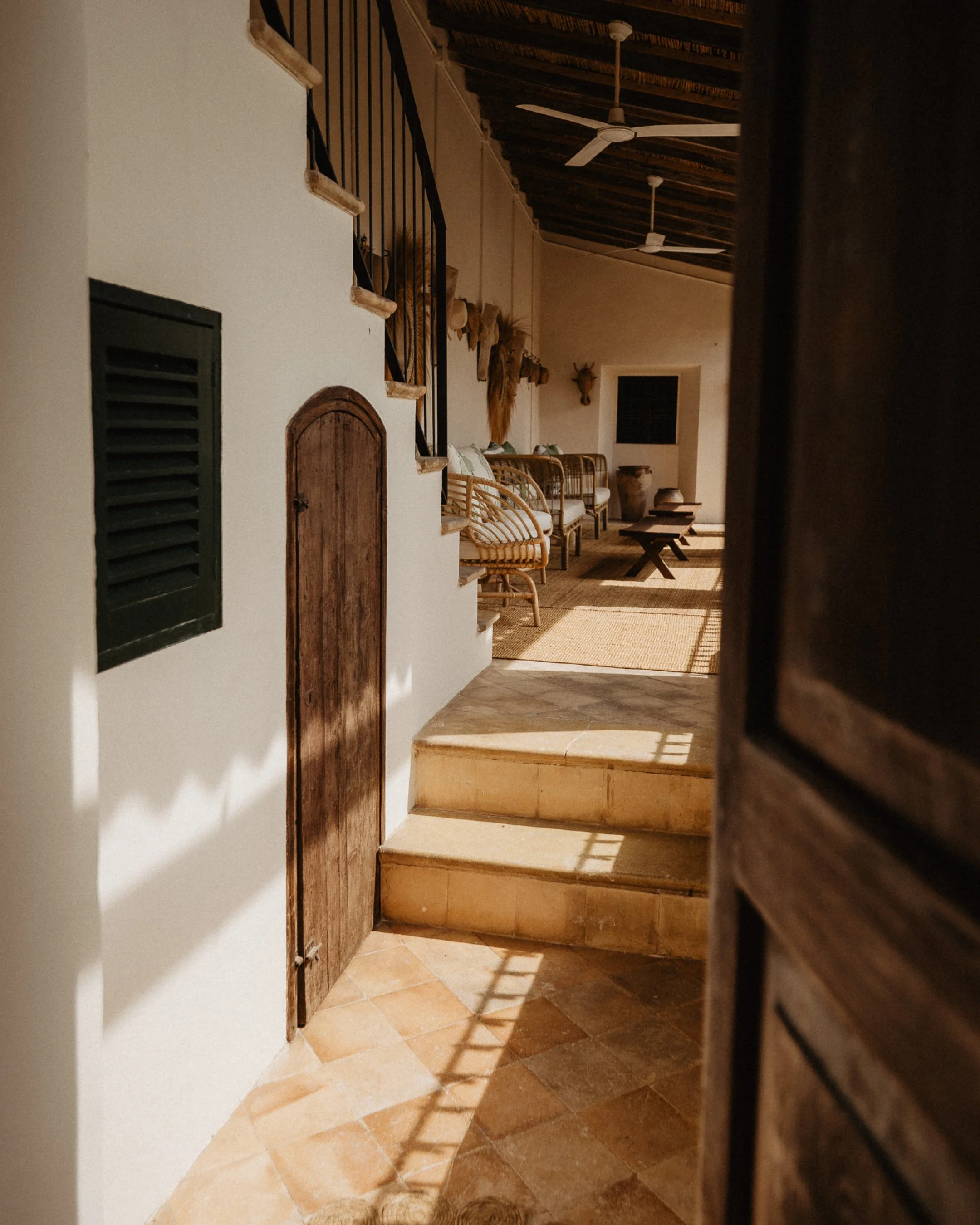 A cozy interior view showing a staircase leading to a sitting area with wicker chairs, wooden coffee tables, and wall-mounted decorations including mounted deer heads and dried plants. Sunlight casts shadows on the tiled floor.