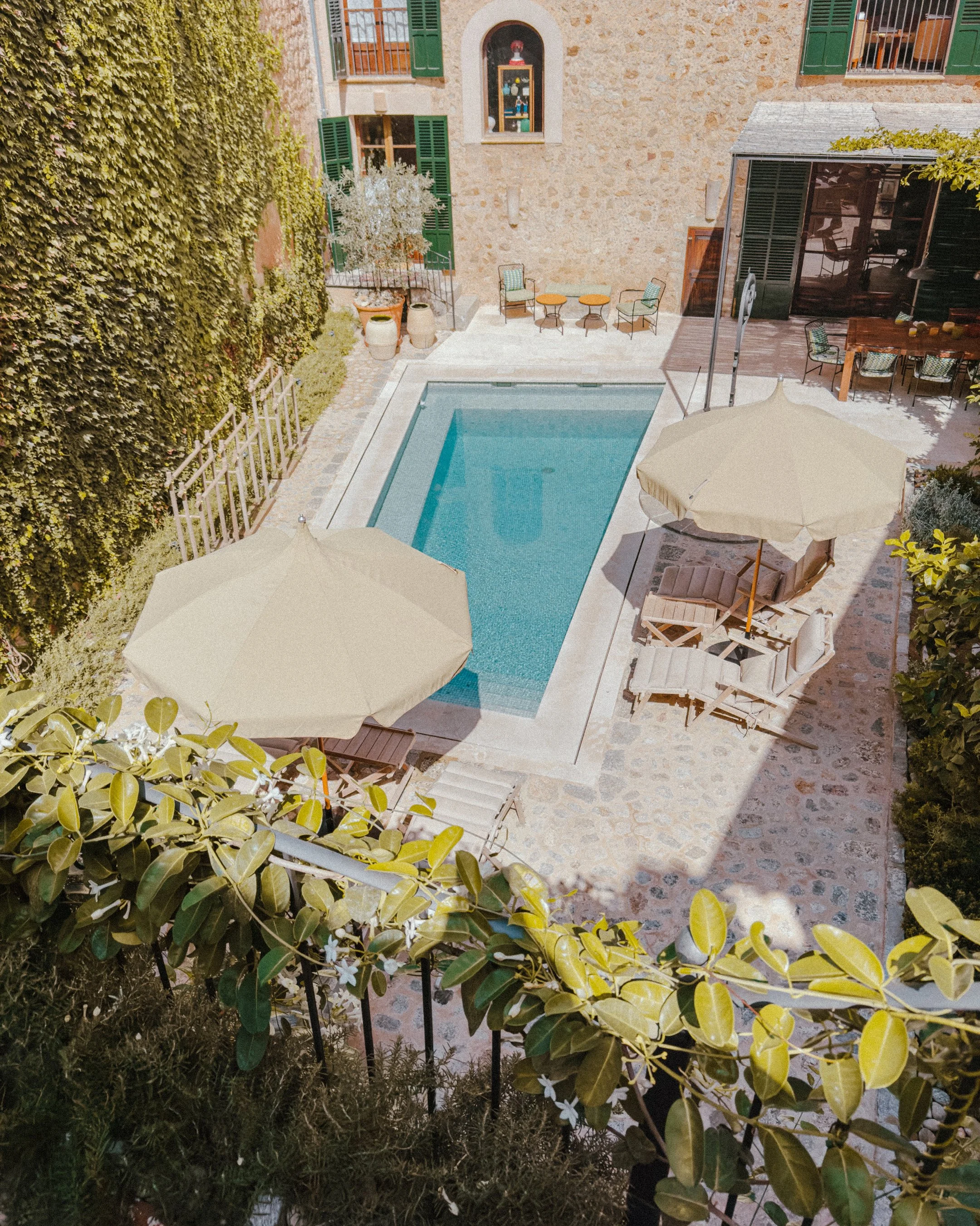 An outdoor pool area with umbrellas, lounge chairs, and a stone building with green shutters.