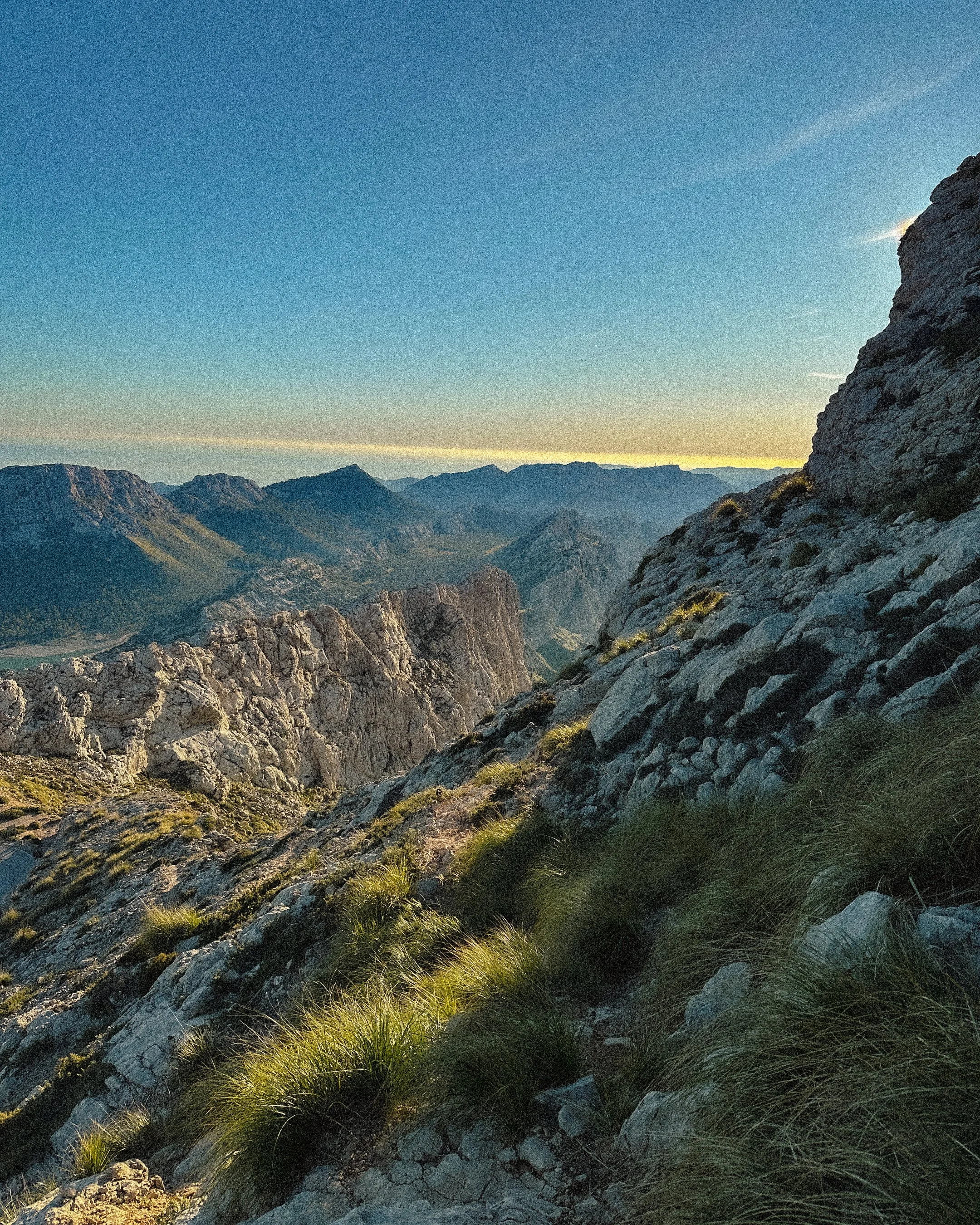 Scenic mountain landscape with rocky slopes, green grass patches, rugged peaks, and a clear blue sky at sunset.