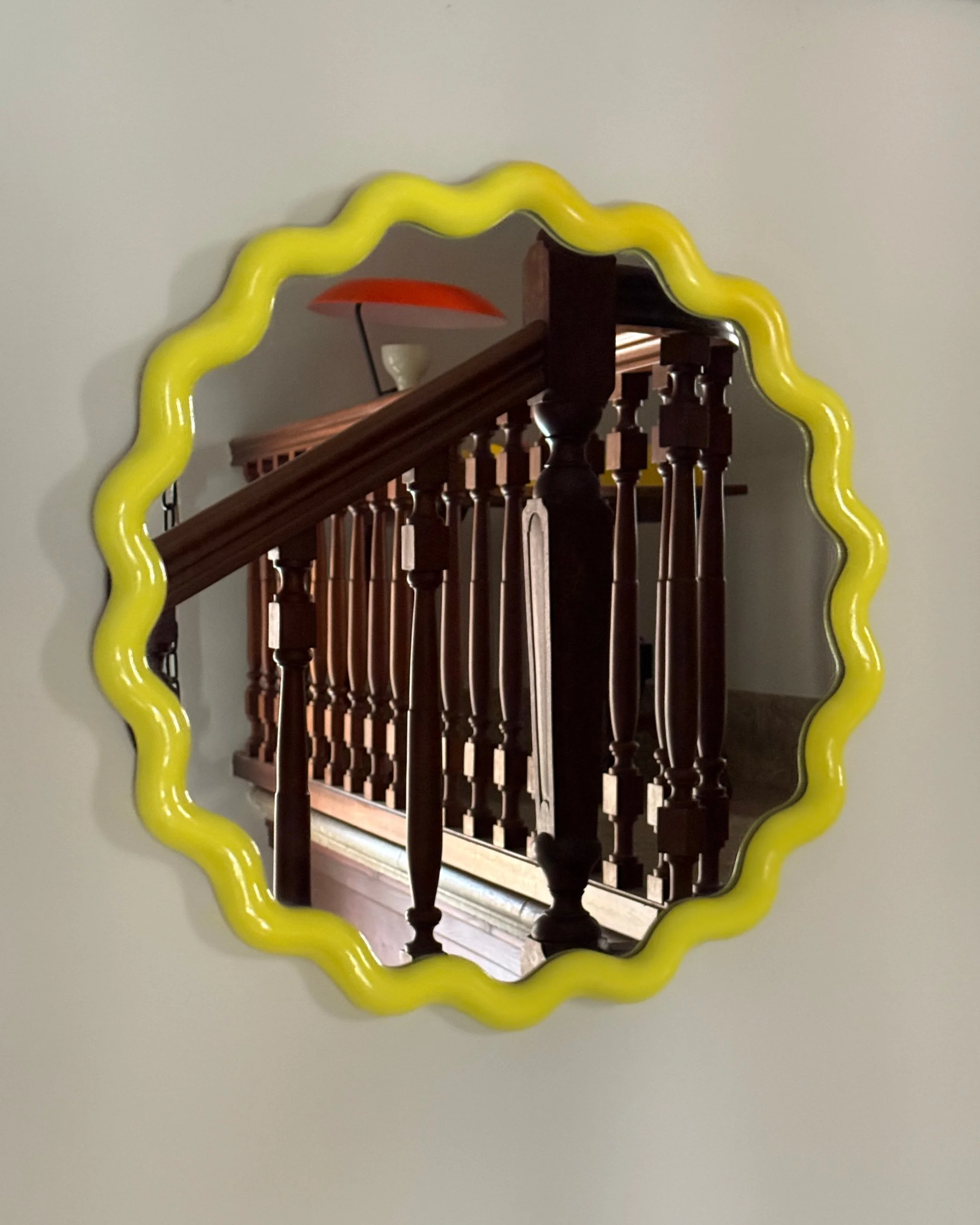 Round mirror with a yellow wavy frame hanging on a wall, reflecting a staircase with wooden banister and spindles, a red wall-mounted lamp, and a small white object on the wall.