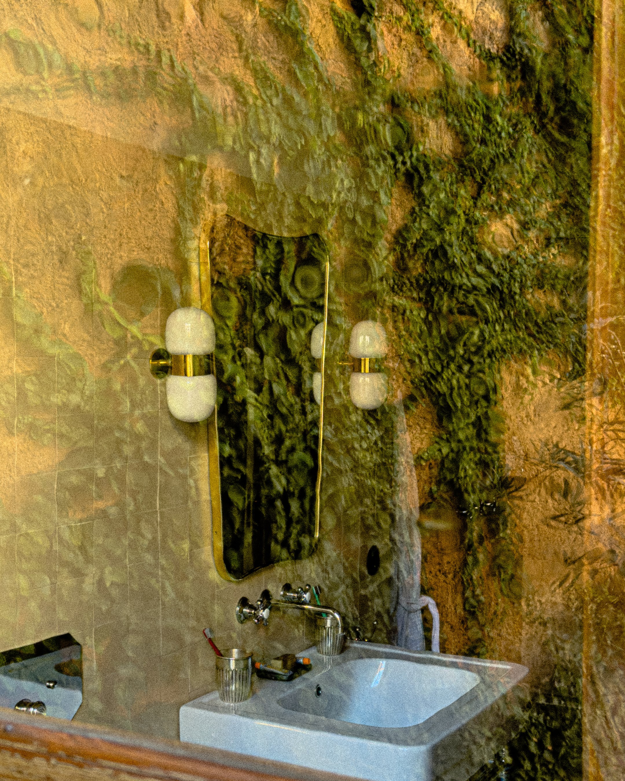 Interior view of a bathroom with a white sink, a mirror, and wall-mounted light fixtures, decorated with green plants and a textured wall design.