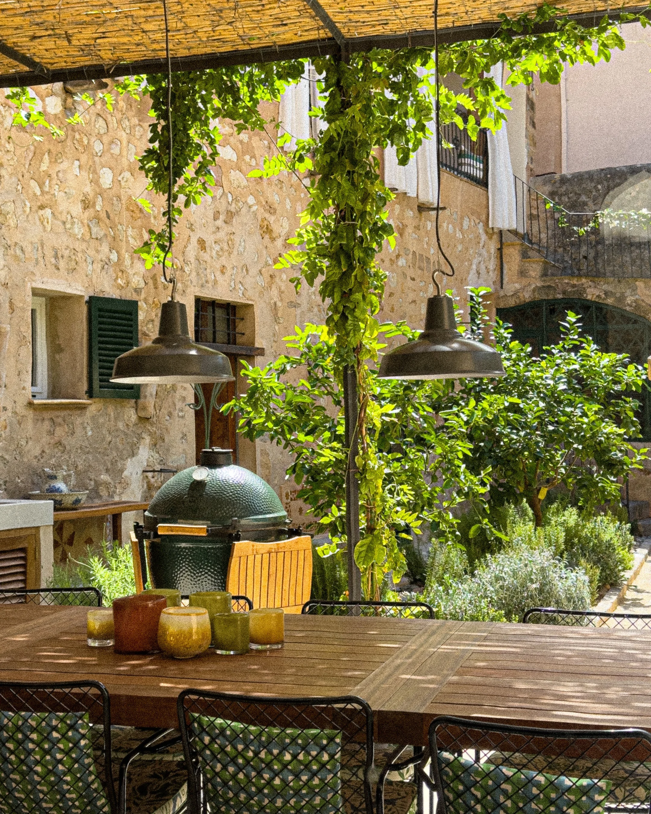 An outdoor patio with a wooden table, six chairs, a large green ceramic grill, and hanging pendant lights, surrounded by greenery and stone buildings with windows and balconies.