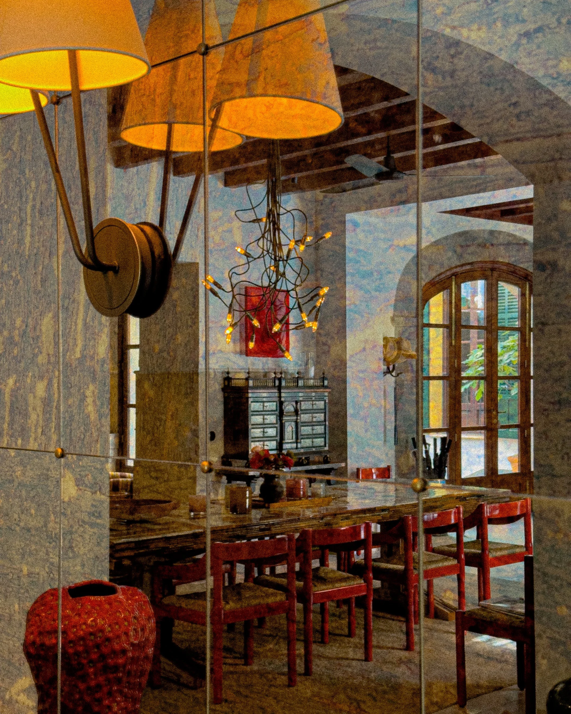 Interior of a dining room with a wooden table and red chairs, a decorative red vase, a vintage cabinet, a large window with wooden shutters, hanging lamps, and a wall mirror reflecting the room.