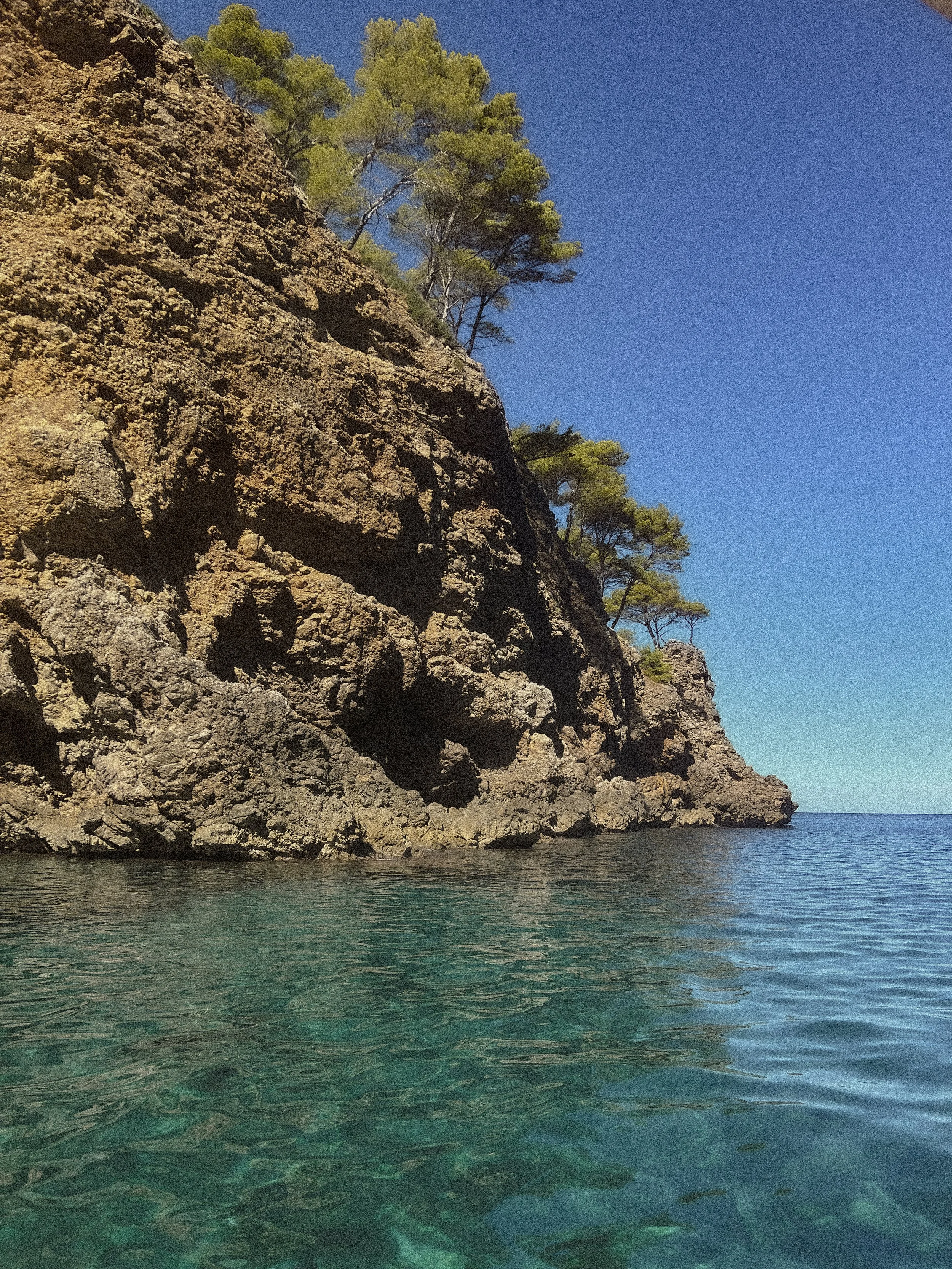 A rocky cliff with green trees on top against a blue sky, and clear turquoise water at the base.