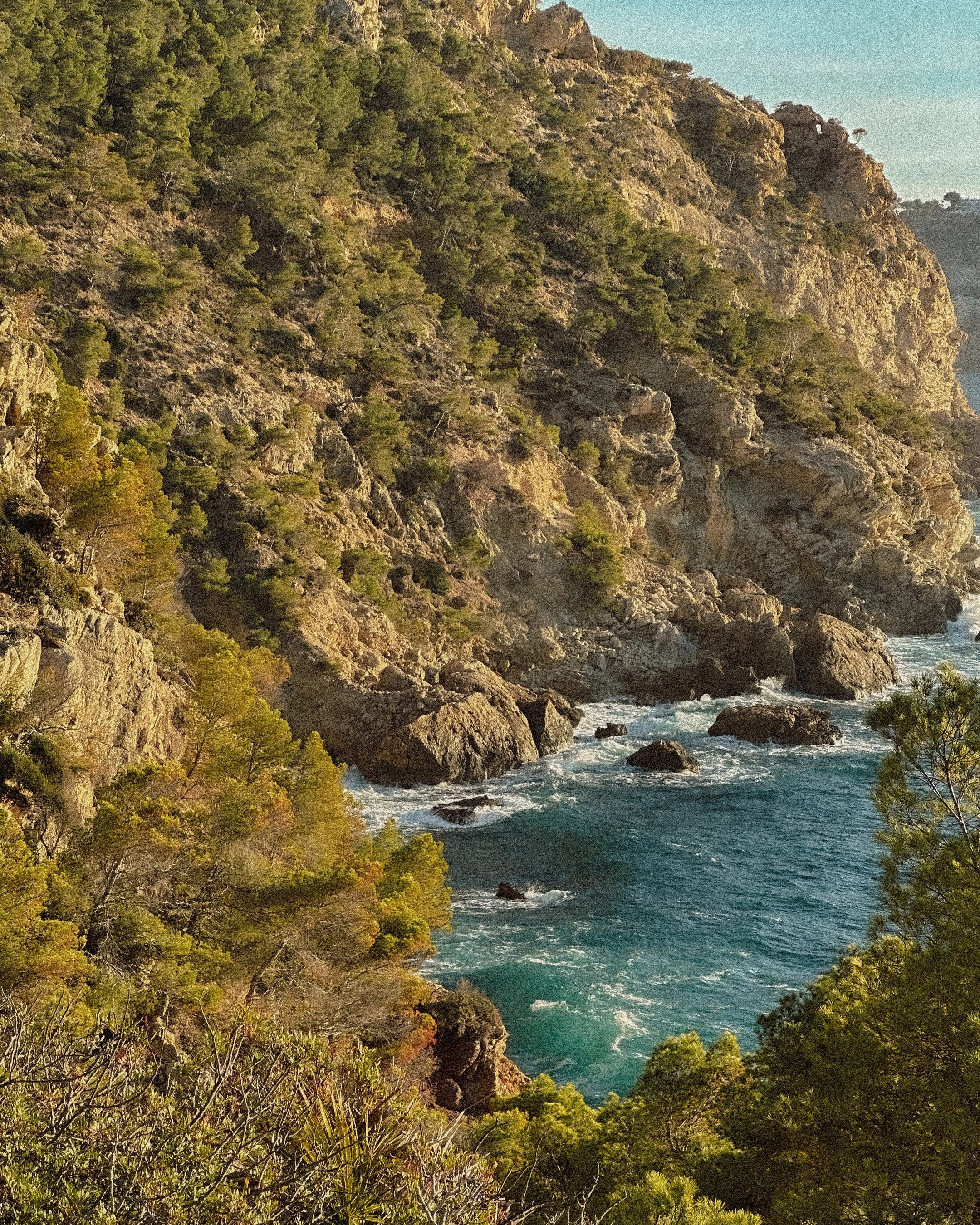 Scenic view of a rugged coastline with rocky cliffs covered in green trees, overlooking the ocean with waves crashing against the rocks.