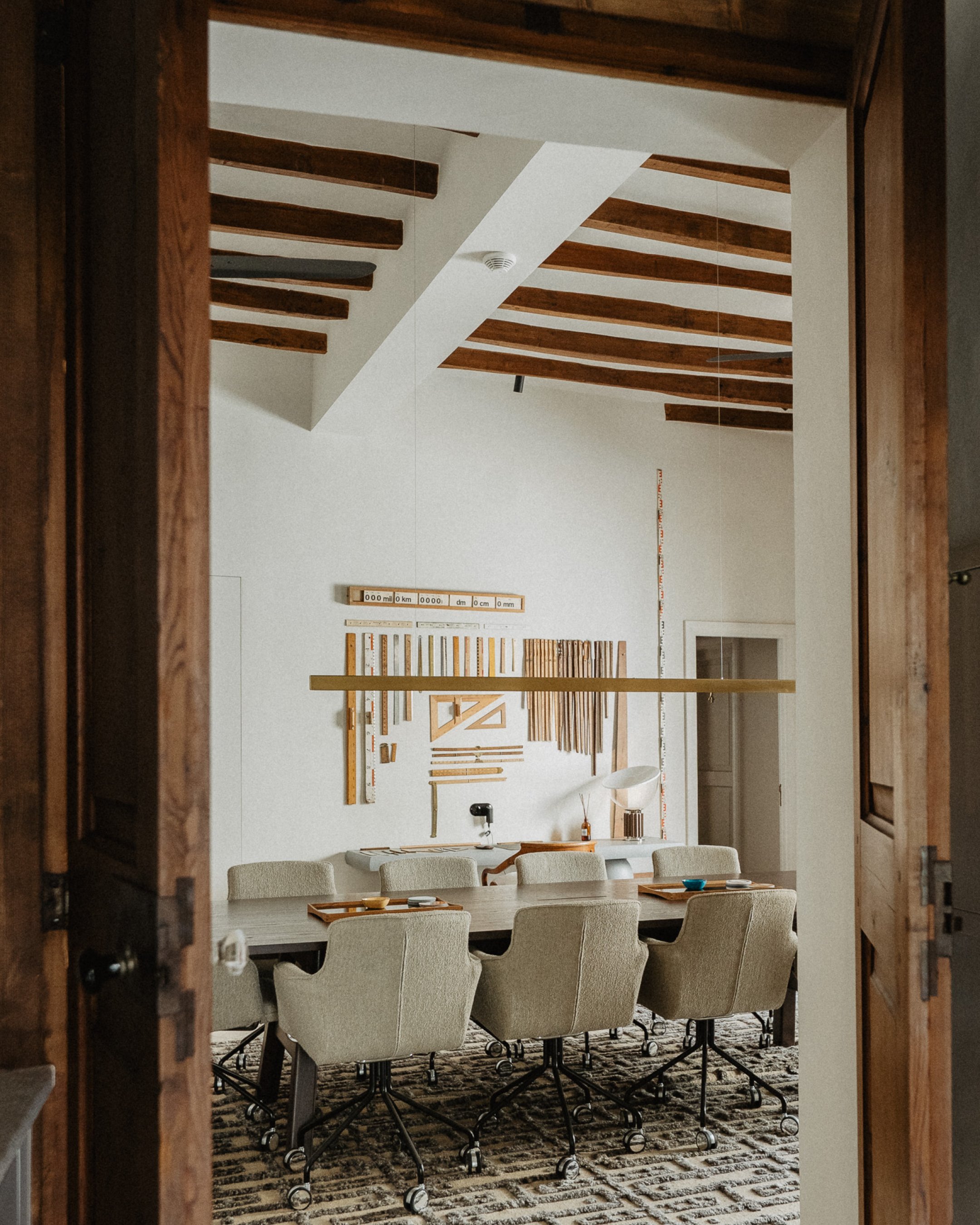 A modern conference room with a wooden table surrounded by beige office chairs on wheels. The room has a textured rug, wooden ceiling beams, and a white wall decorated with wooden rulers and measuring tools. There is a small desk with a lamp and some items on it.