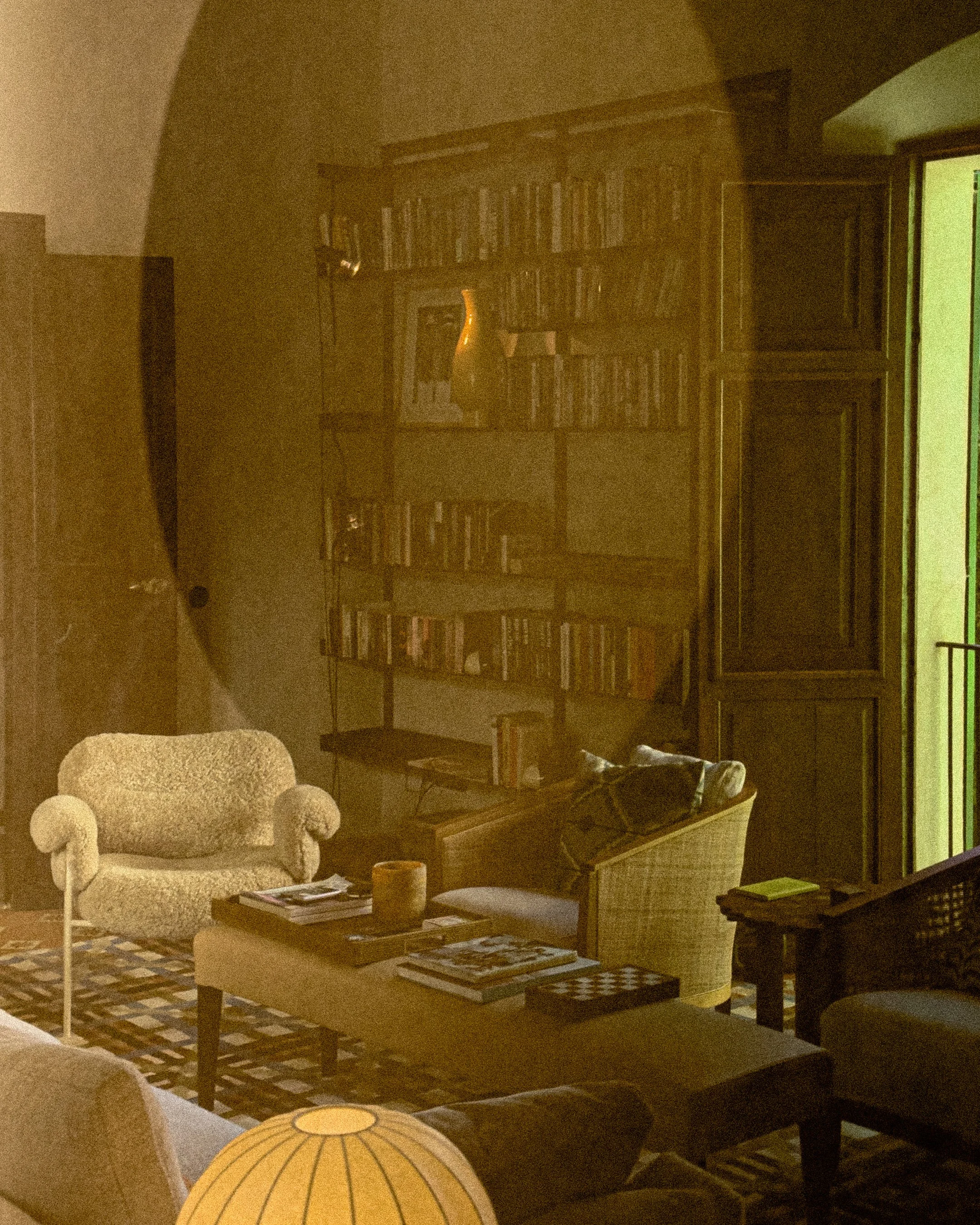 Living room with beige armchair, coffee table with magazines, bookshelf filled with books, wooden cabinet, and slippers on a chair. Dim lighting with a textured rug.