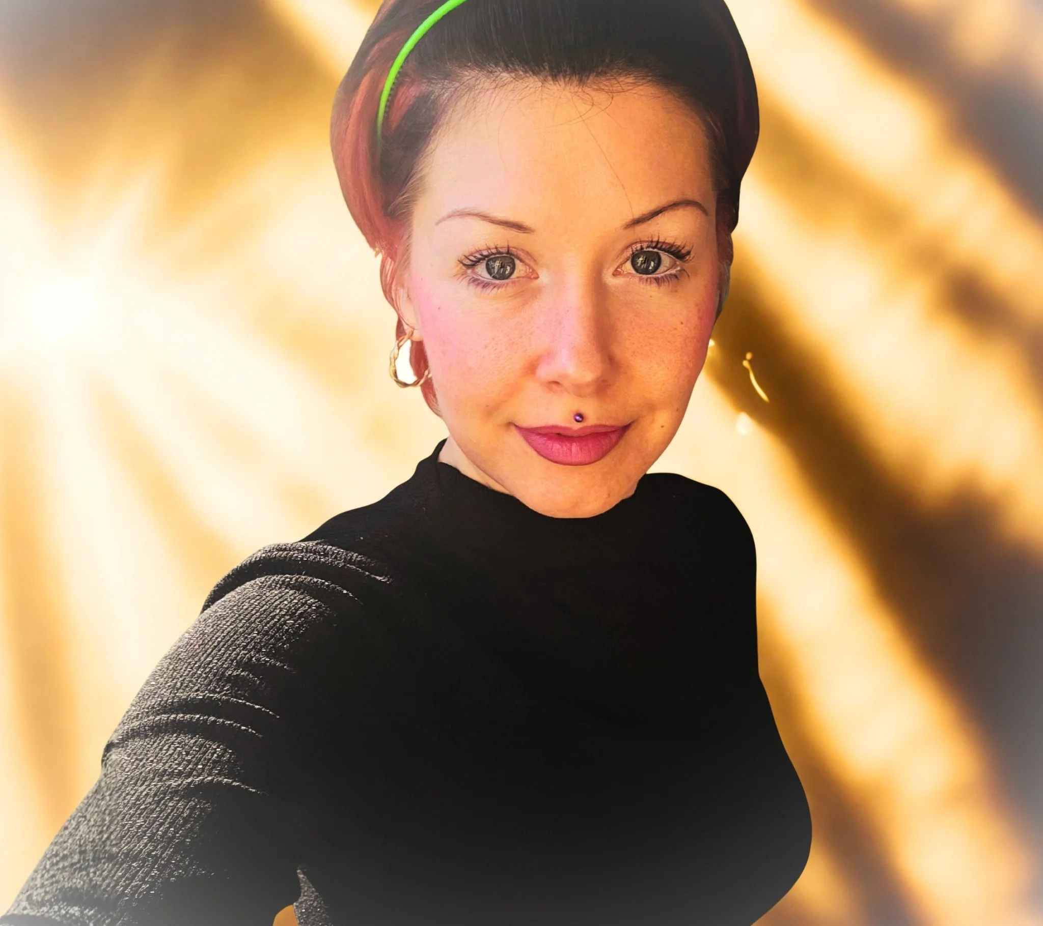 Close-up of a woman with short hair, bright eyes, and multiple facial piercings, wearing a black top and gold hoop earrings, with a blurred gold and black background.