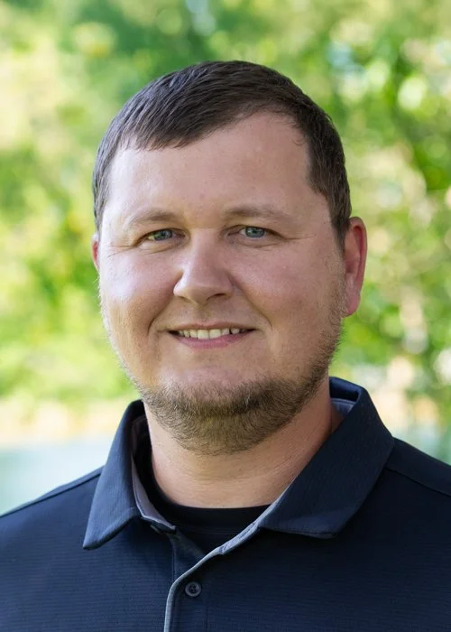 A young man with short brown hair and a beard, wearing a dark collared shirt, smiling outdoors with green trees in the background.