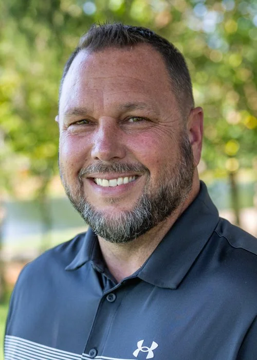 A smiling man with a beard and short dark hair wearing a black Under Armour polo shirt outdoors with blurred green trees in the background.