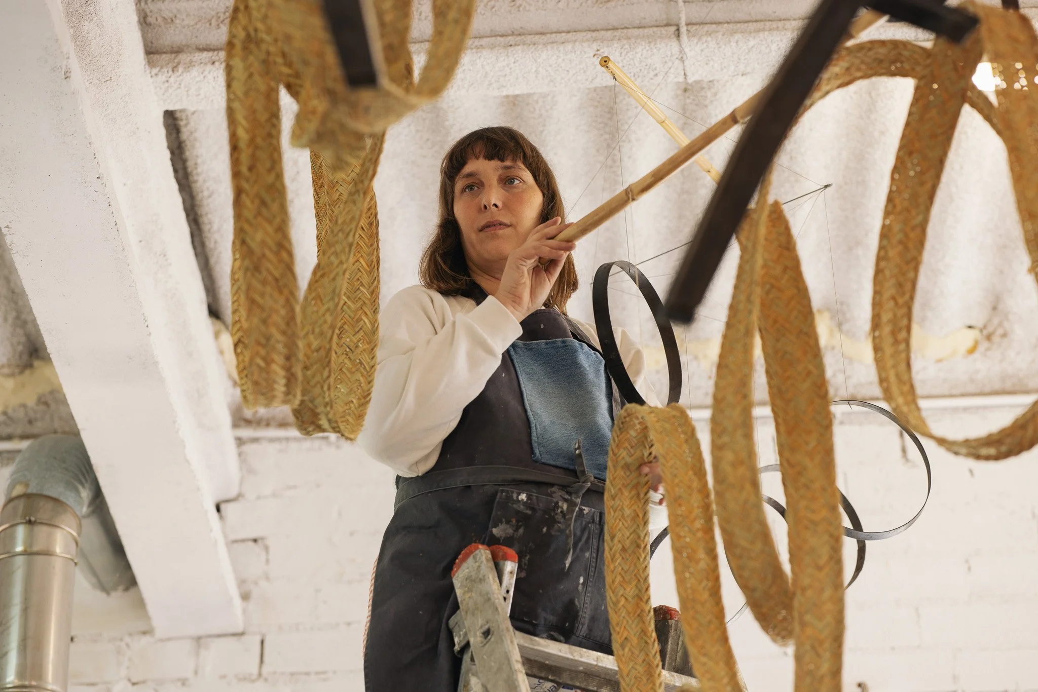 A woman standing on a ladder, hanging woven ropes or decorations from a ceiling with a wooden stick, in a room with white brick walls.