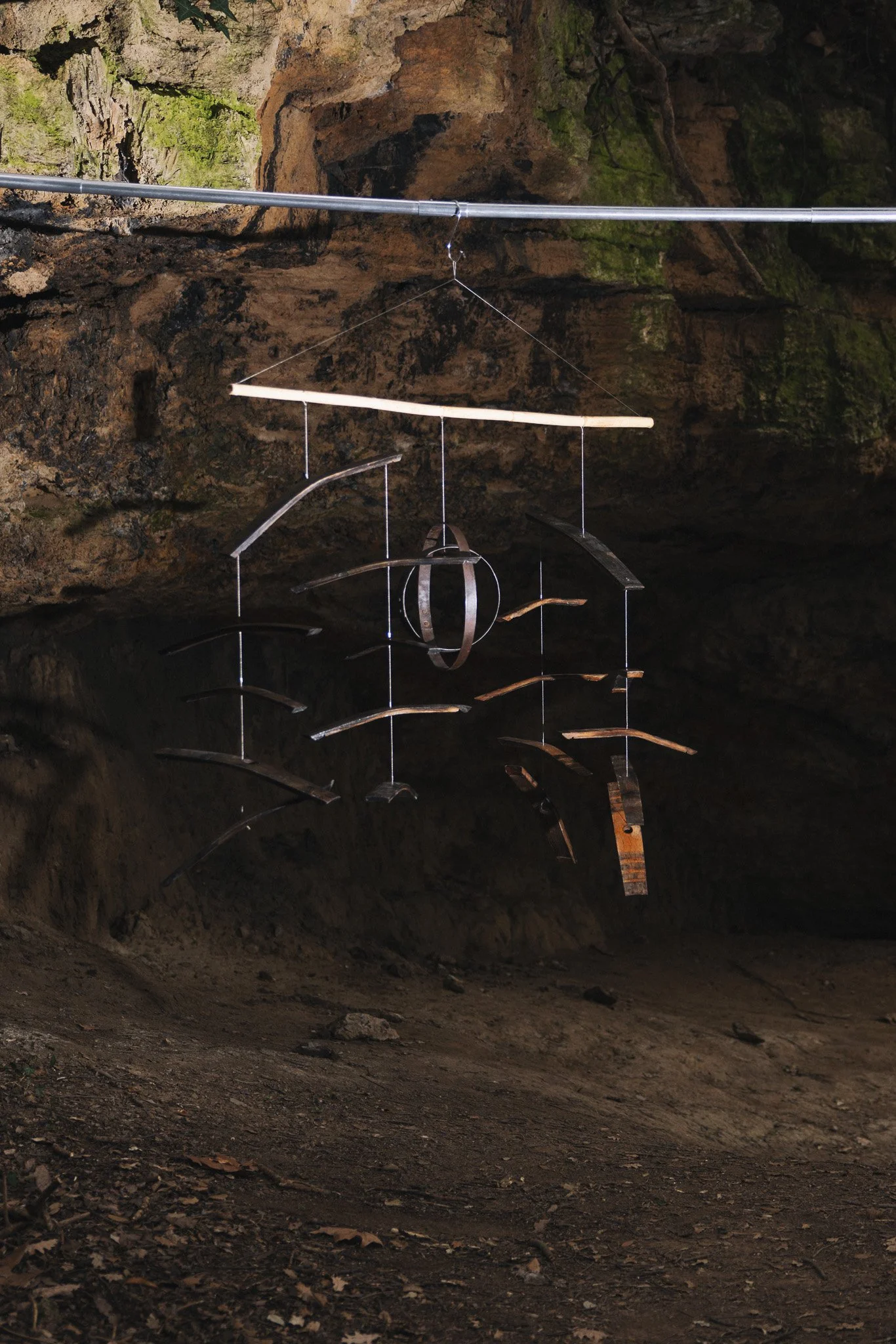Metal wind chimes hanging from a metal rod inside a cave with mossy rocks.