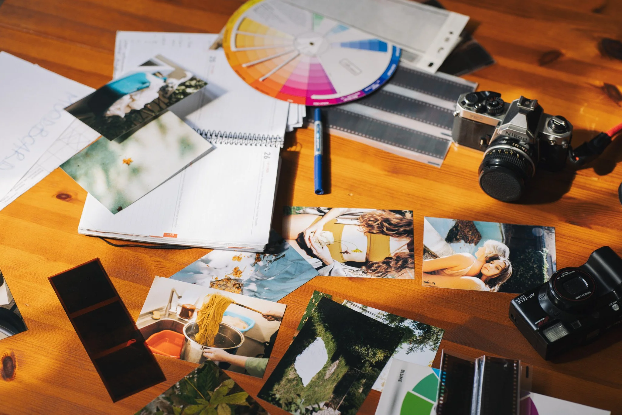 A wooden table covered with photography and design materials, including printed photographs, a color wheel, film strips, a camera, a notebook, and a color palette.