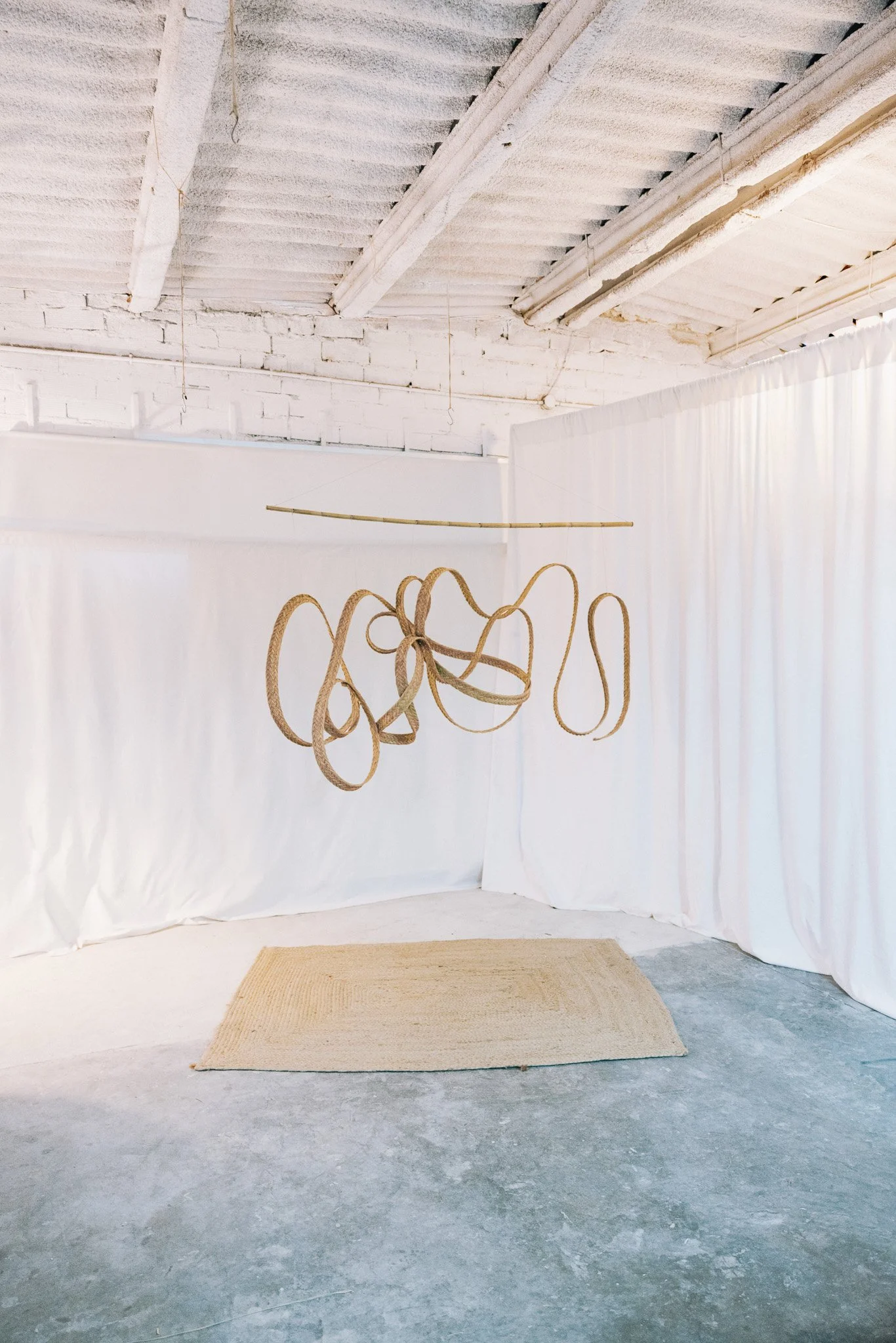 Art installation with a hanging sculpture made of intertwined beige ropes, suspended from a curved rod in a minimalist gallery space with white curtains and a beige rug on a concrete floor.