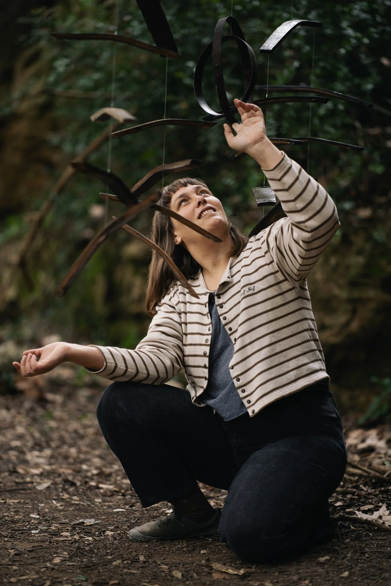 Woman in striped jacket crouching outdoors, reaching up towards a hanging metallic sculpture.