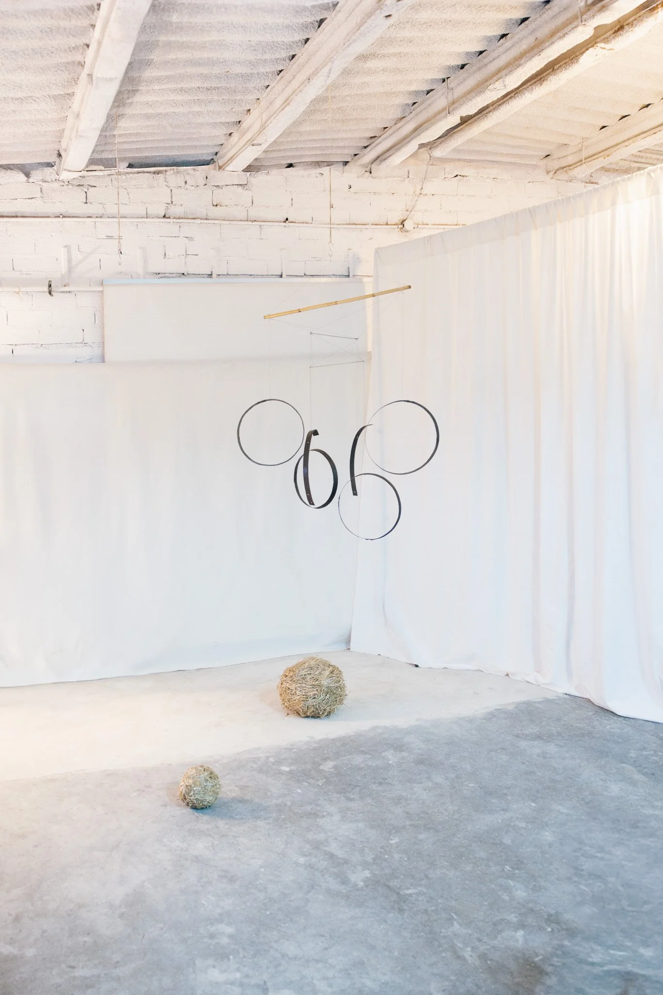 Minimalist art installation with hanging circles and numbers, and two textured spherical objects on the floor, set against white curtains and a white brick wall in an industrial space.