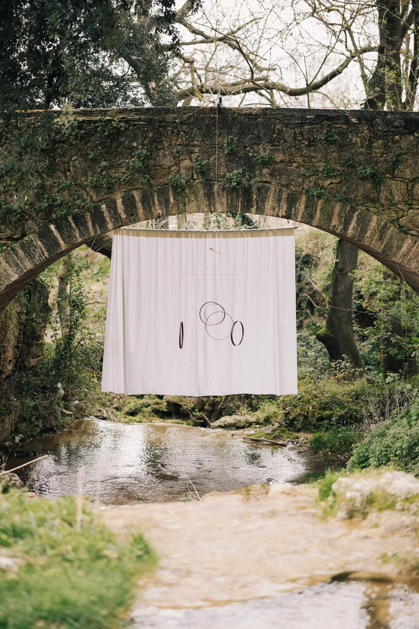 A white curtain with black circular designs hanging from a bridge over a creek in a wooded area.