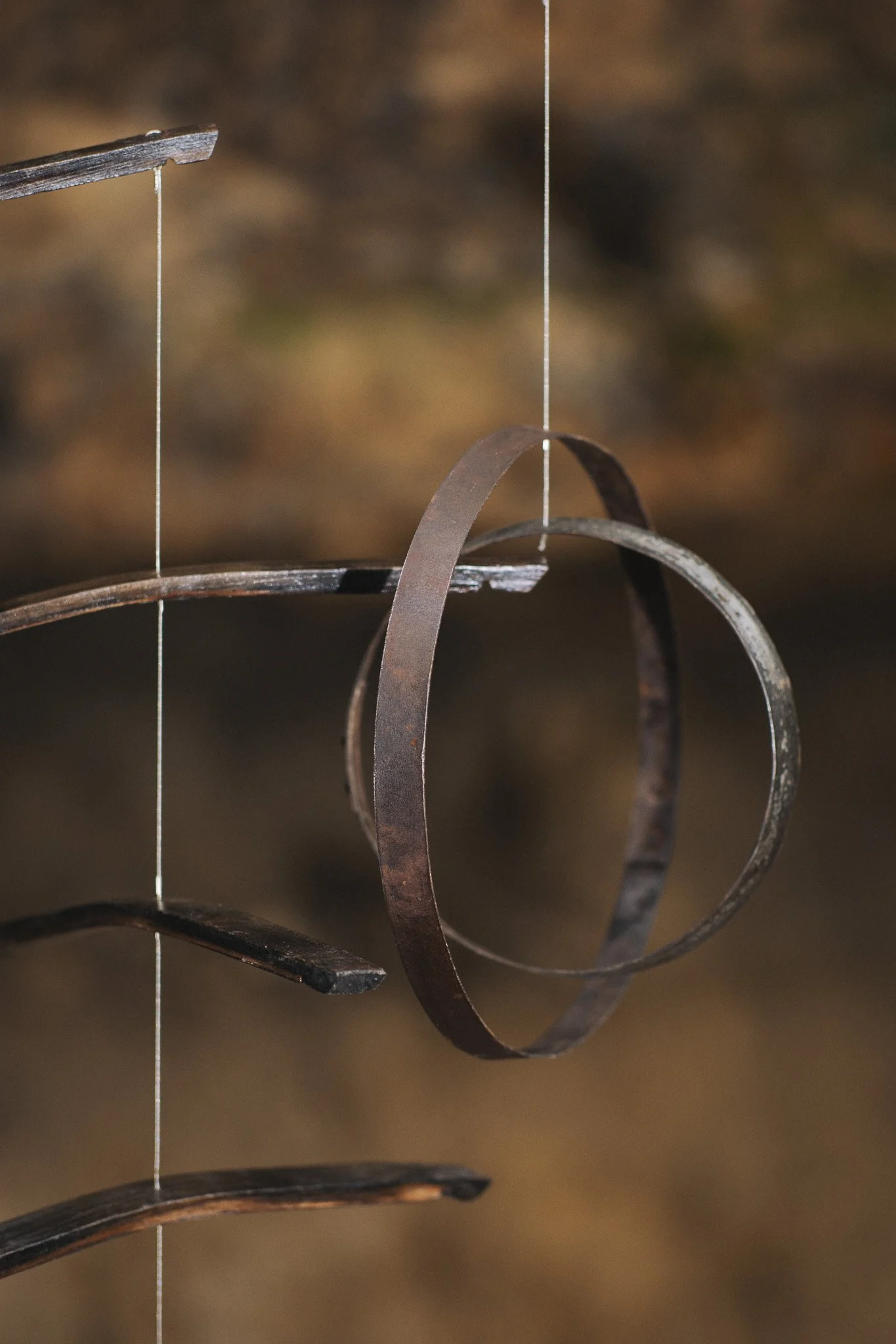 Close-up of a metal mobile sculpture with circular and curved metallic pieces hanging from thin wires, against a blurred brown background.