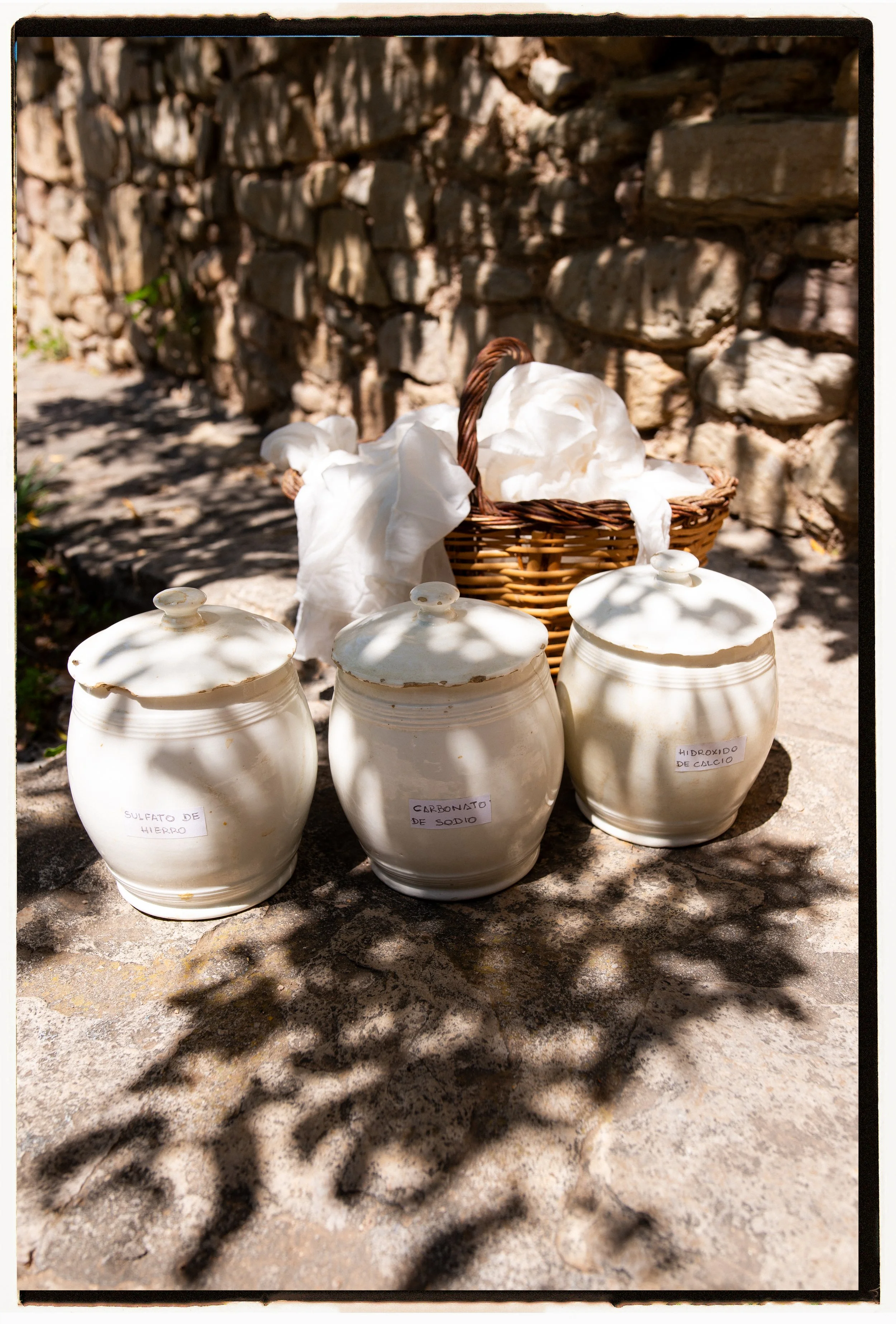 Three white ceramic jars with labeled lids, a wicker basket filled with white cloth or tissue paper, placed outdoors on a stone surface with a stone wall in the background, and shadows of tree leaves cast on the ground.
