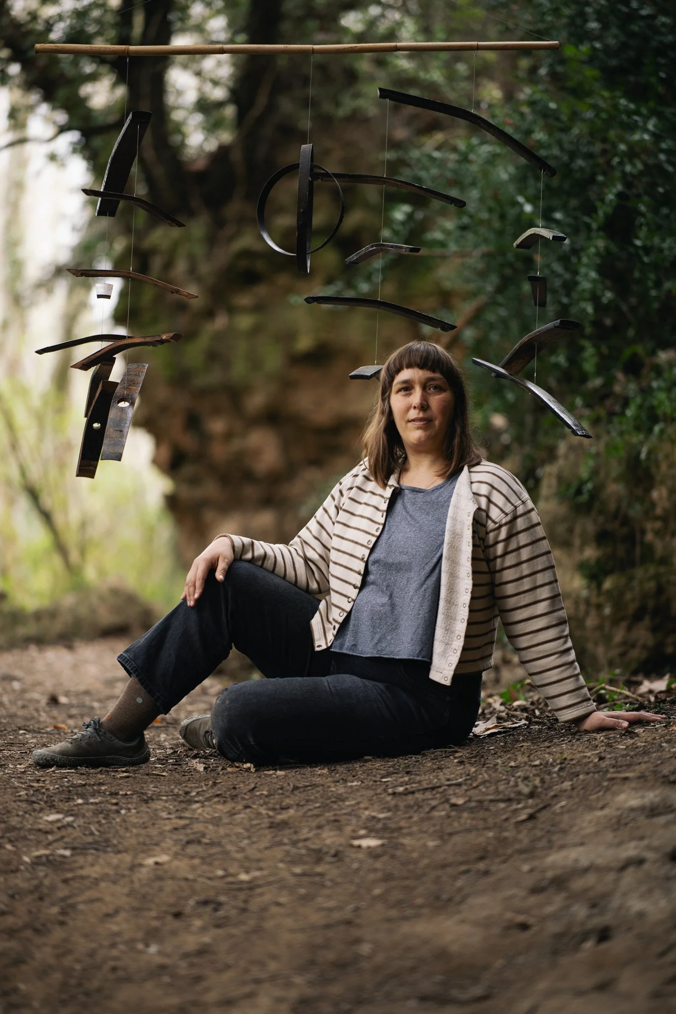 A woman sitting on the ground in a forest with a wind chime hanging above her.