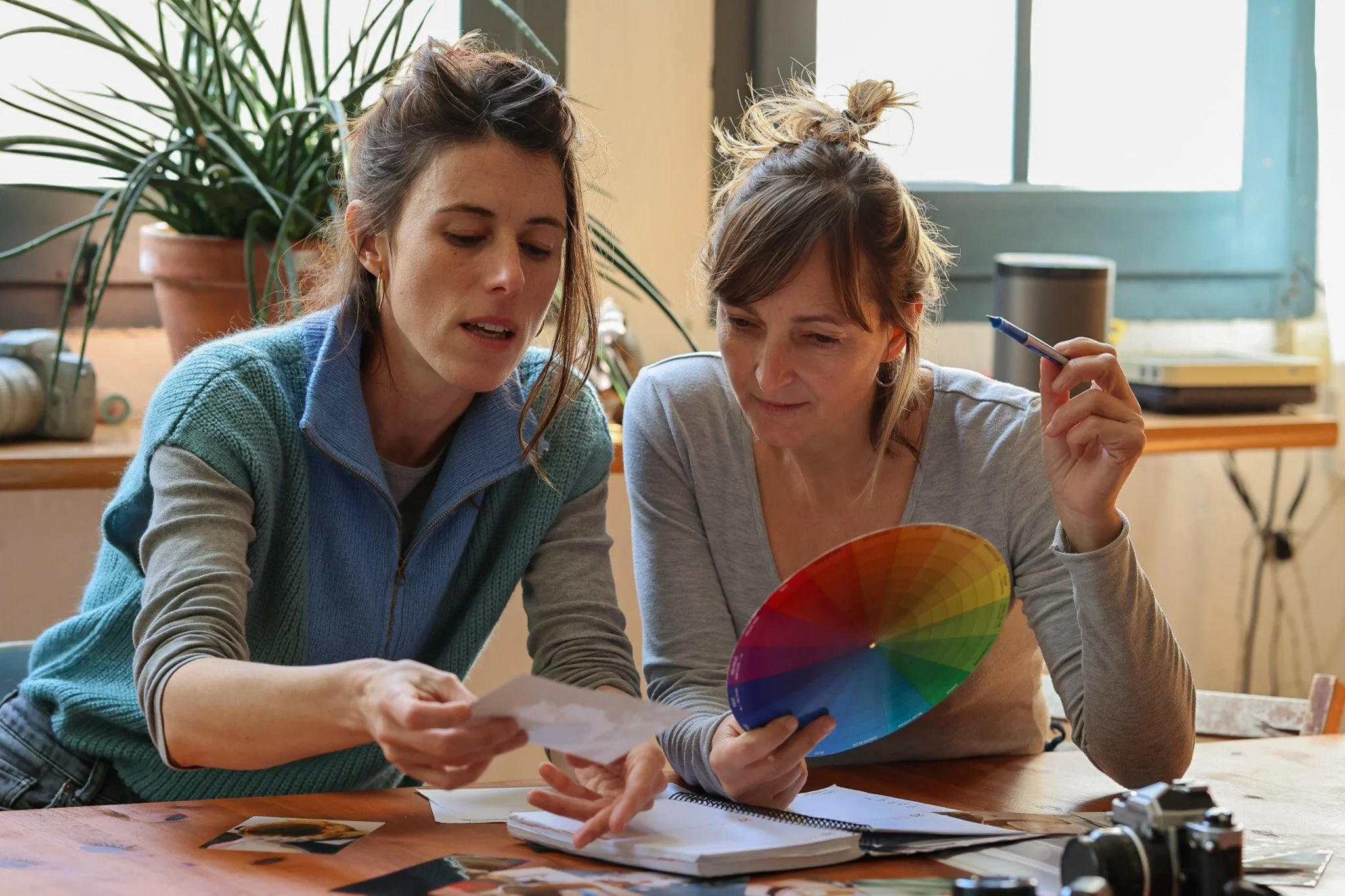 Two women are sitting at a wooden table, discussing photos and color swatches. One is holding a color wheel and a pen, and the other is holding a photograph, with photographs and papers spread out on the table. There is a large potted plant behind them and a window with turquoise framing in the background.