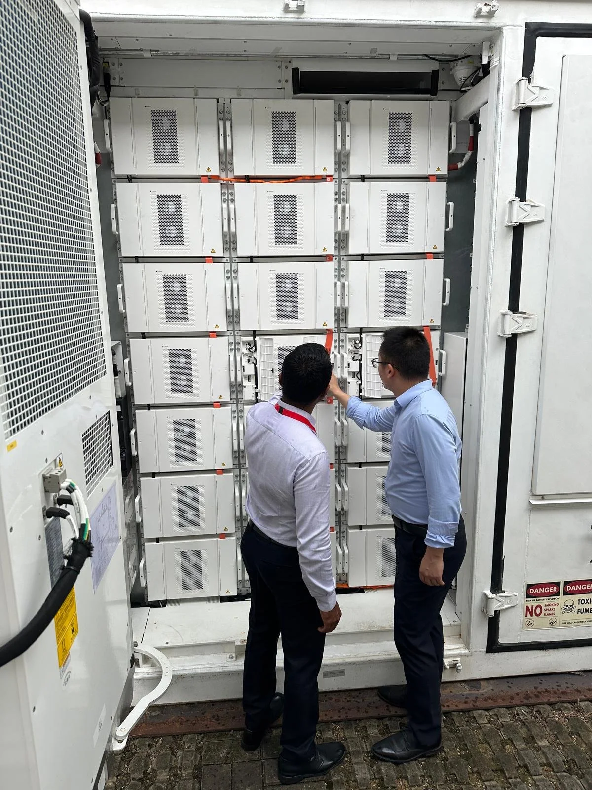 Two men inspecting or working on electrical equipment inside a large industrial electrical cabinet or server enclosure.