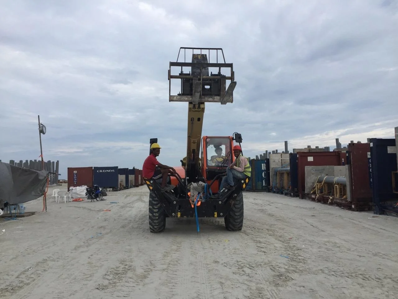 Construction site with two workers wearing helmets, sitting on a piece of construction equipment, with shipping containers and cloudy sky in the background.