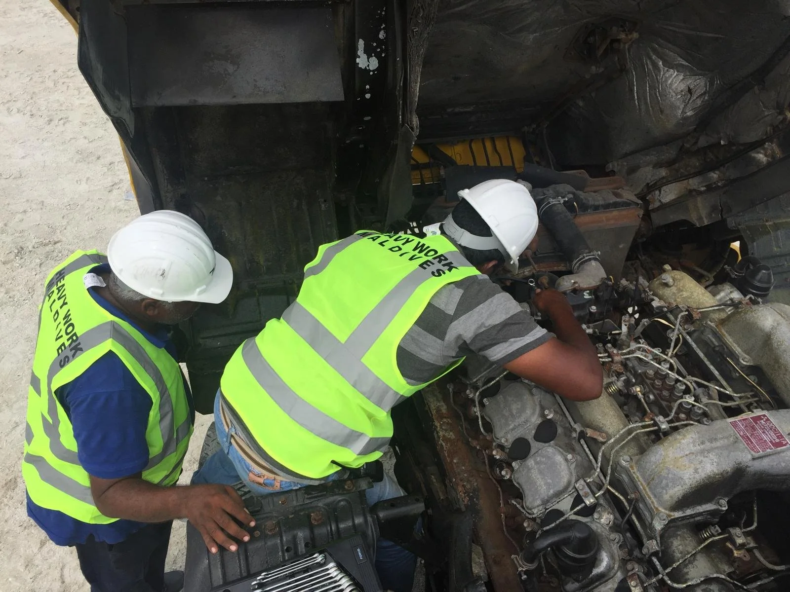 Two workers wearing safety vests and helmets inspecting the engine of a large vehicle.