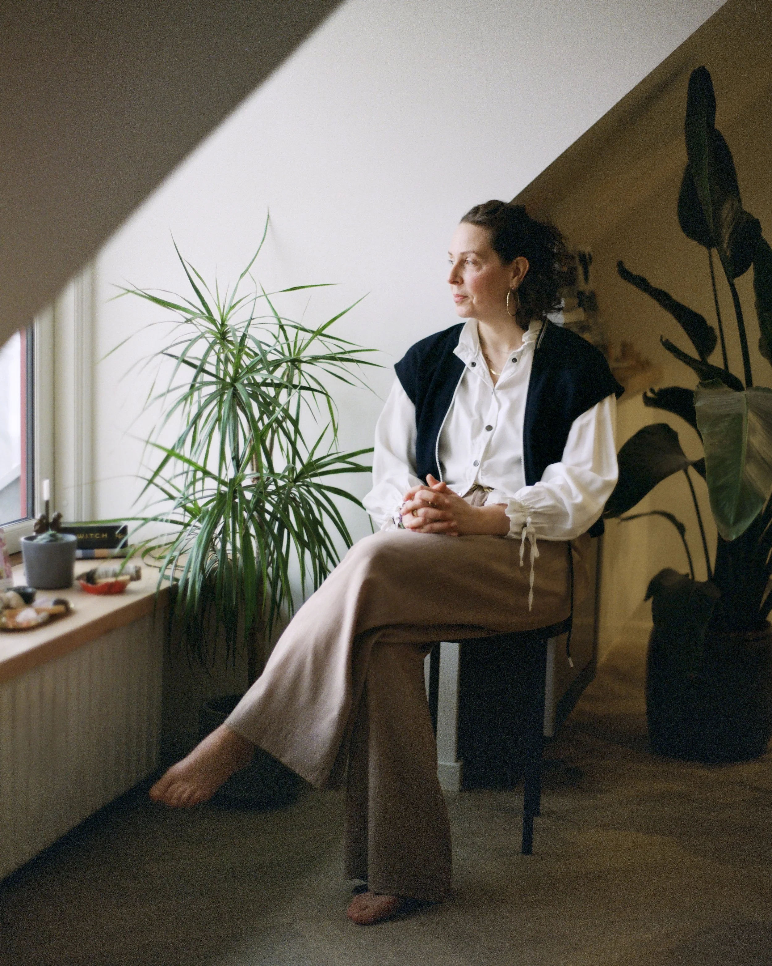 A woman sitting on a chair near a window with potted plants on the windowsill, looking outside thoughtfully.