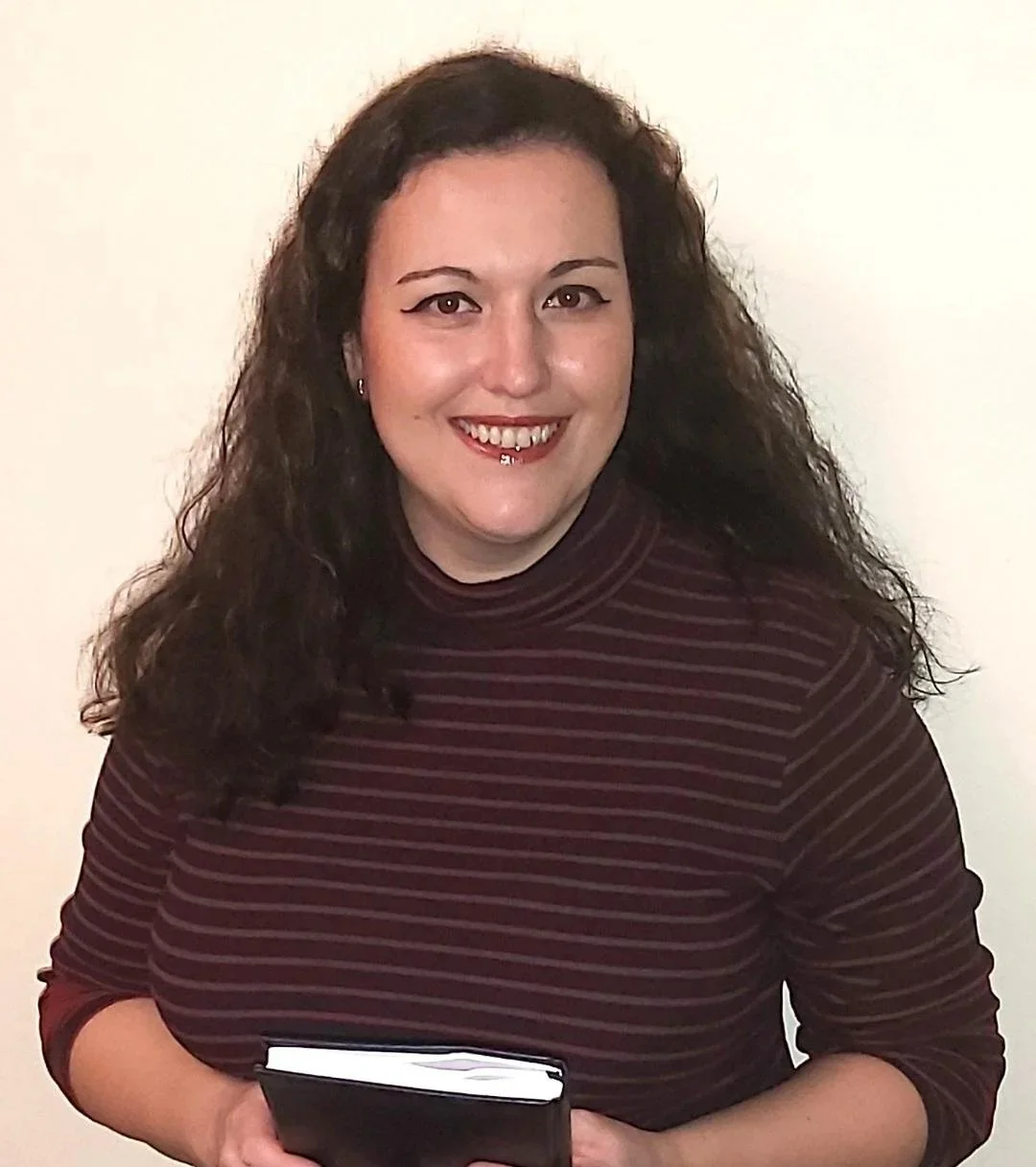 A woman with long curly dark hair wearing a maroon and black striped turtleneck holding a closed book, smiling in front of a plain light-colored wall.