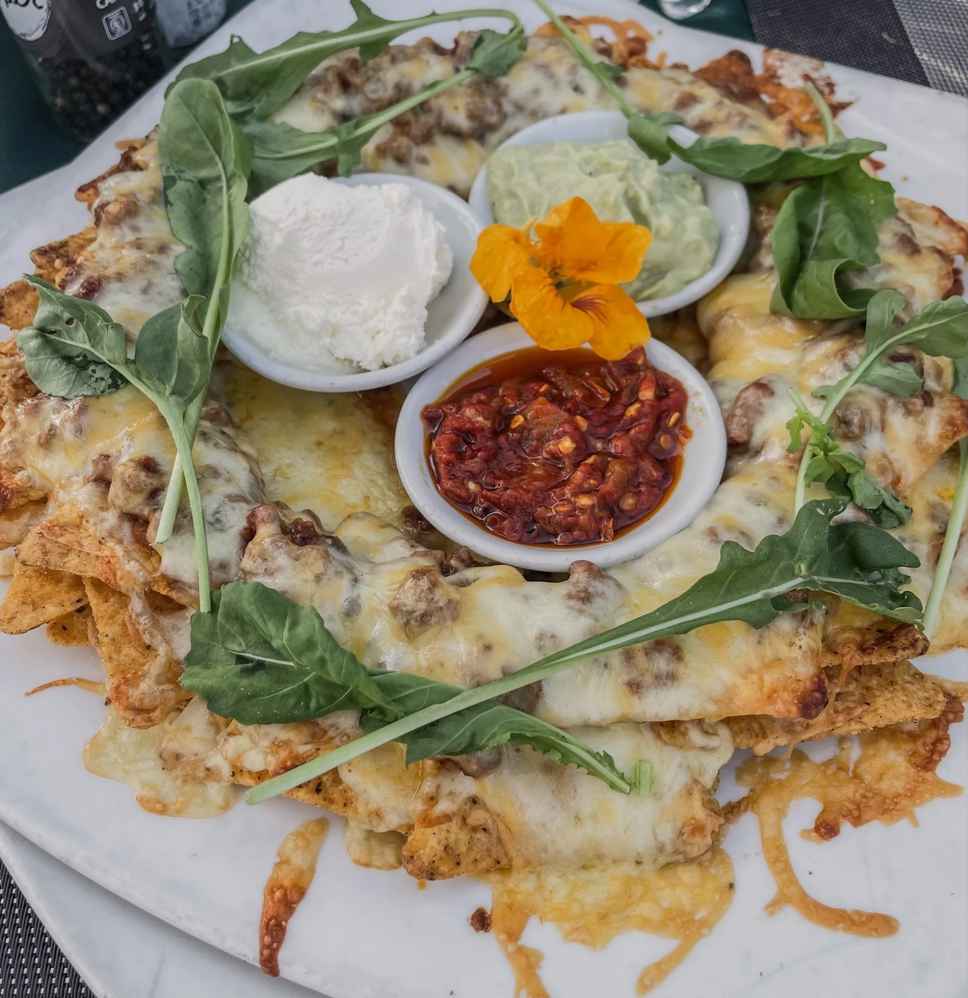 Nachos met gesmolten kaas en drie dips in schaaltjes, met rucola erbovenop. Op terras in Zuid-Afrika.