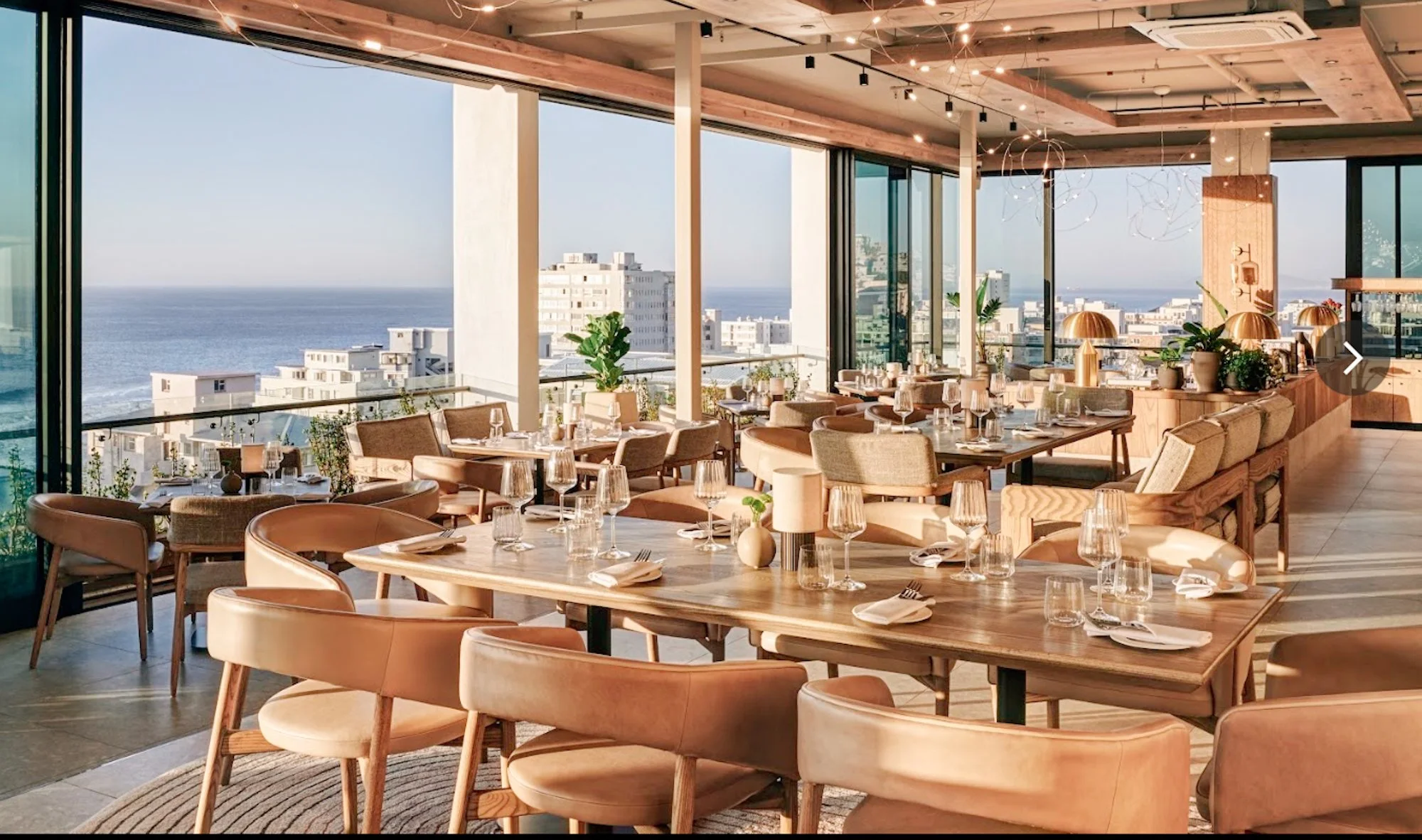 Licht restaurantinterieur van The Nines met houten meubels en uitzicht over Sea Point en de oceaan.