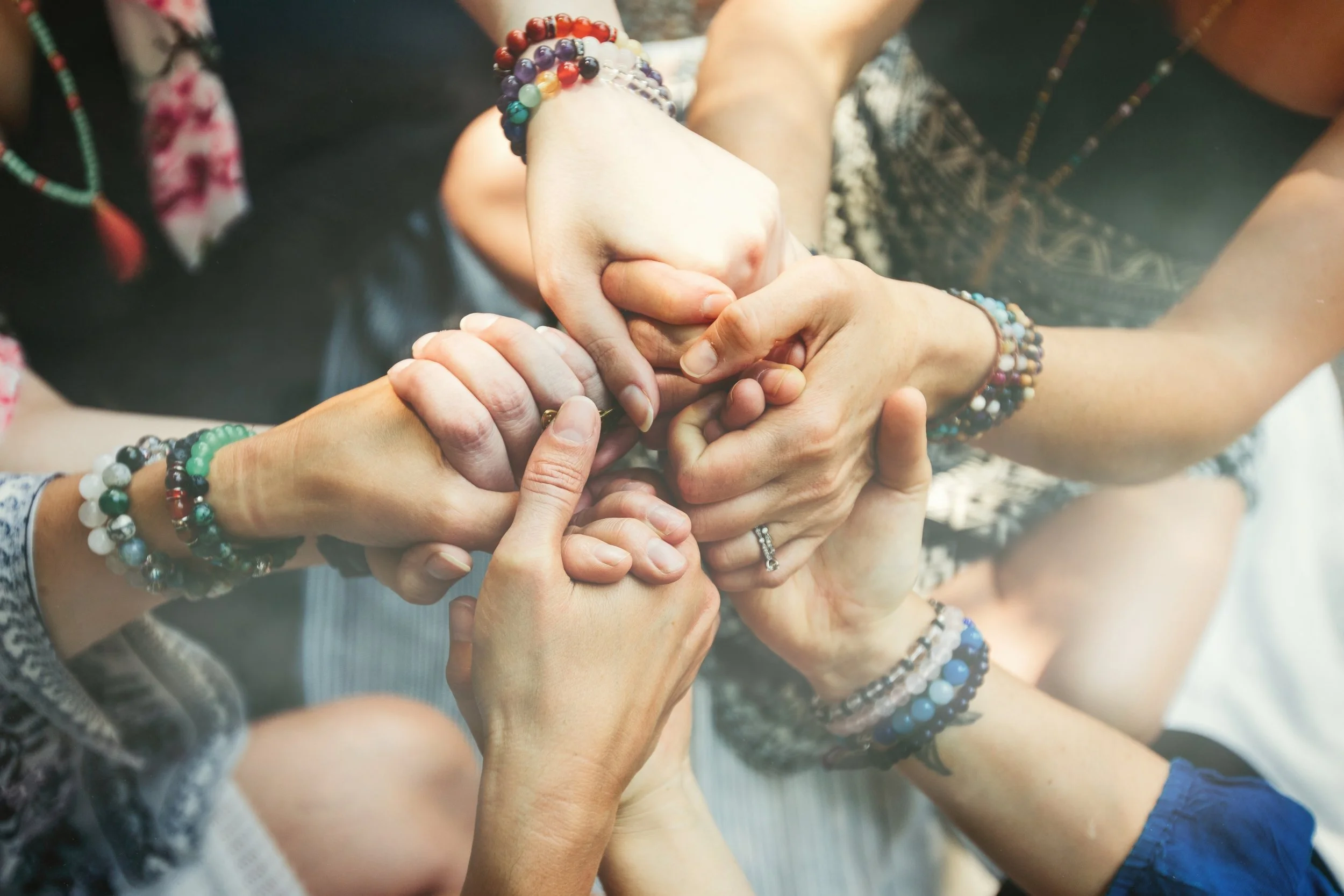 Close-up of multiple hands holding and supporting each other in the center, wearing colorful beaded bracelets and rings, suggesting unity and support.