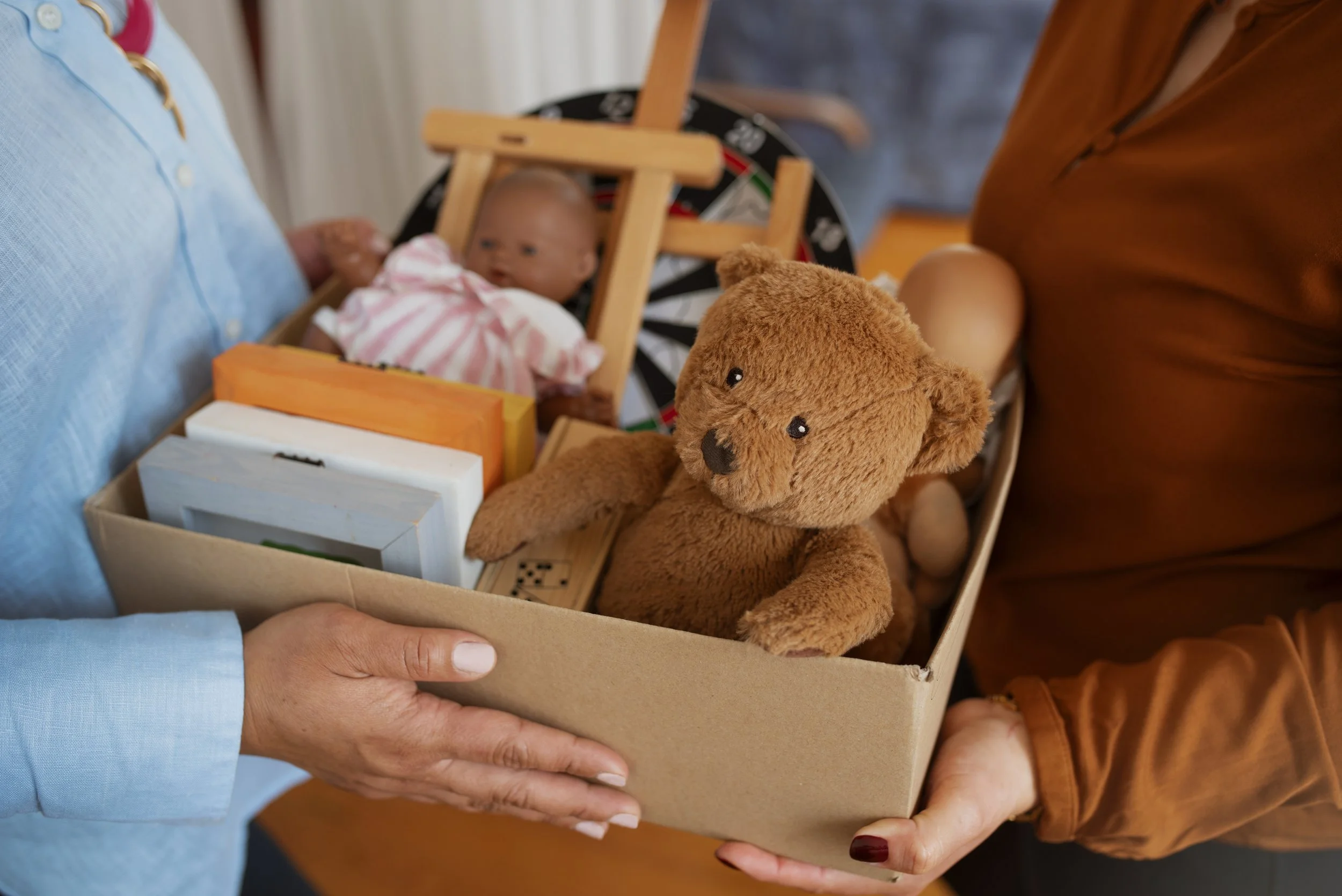 Person holding a cardboard box filled with books, a teddy bear, and a baby doll, with a baby in a high chair in the background.