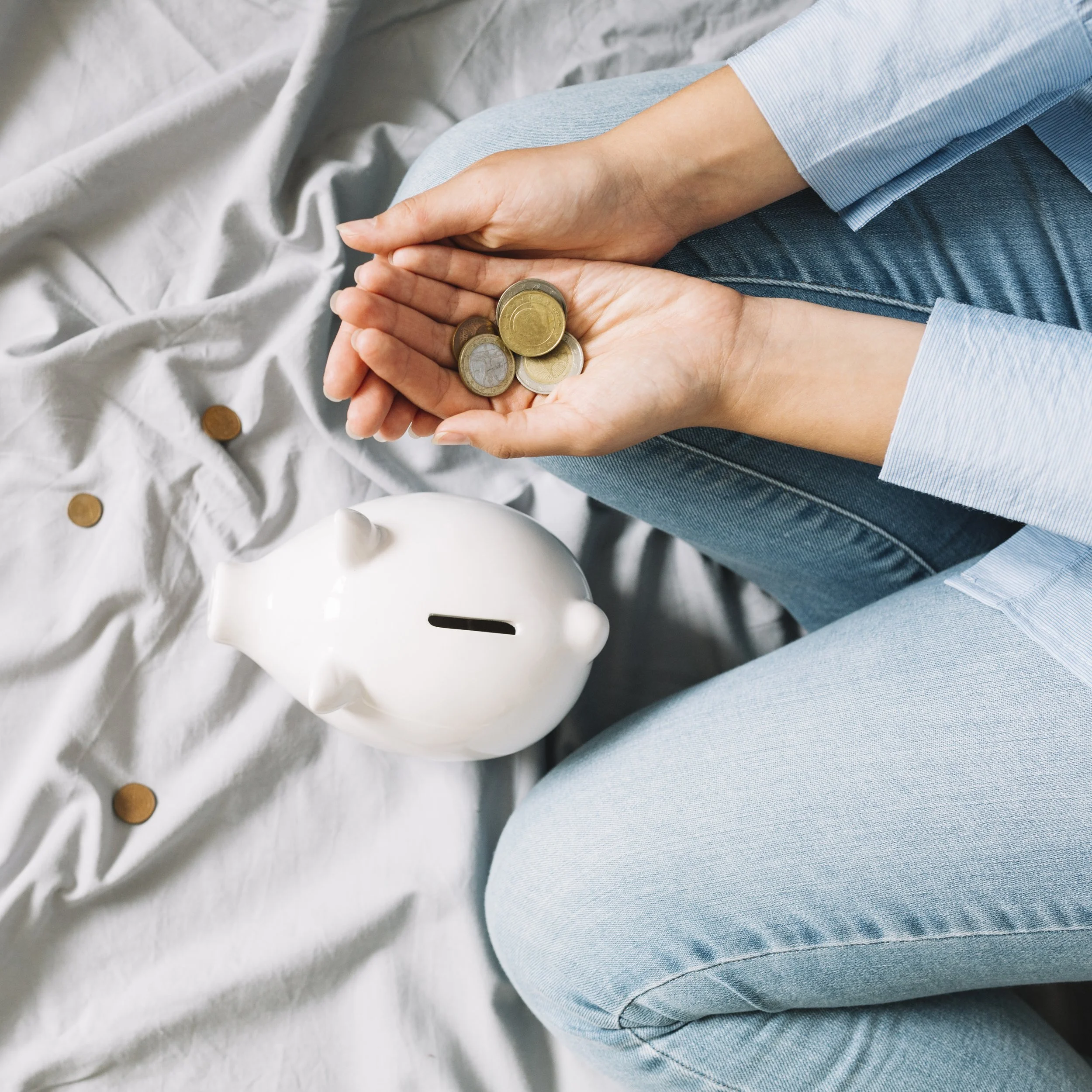 Person sitting on bed holding euro coins in hand, piggy bank visible nearby.