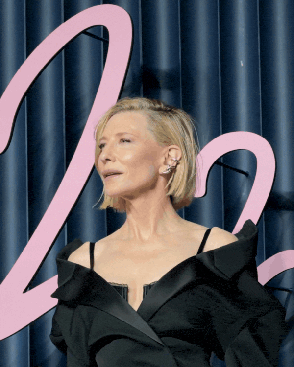 A woman with short blonde hair and multiple earrings poses in front of a dark blue curtain and a pink artistic sign.